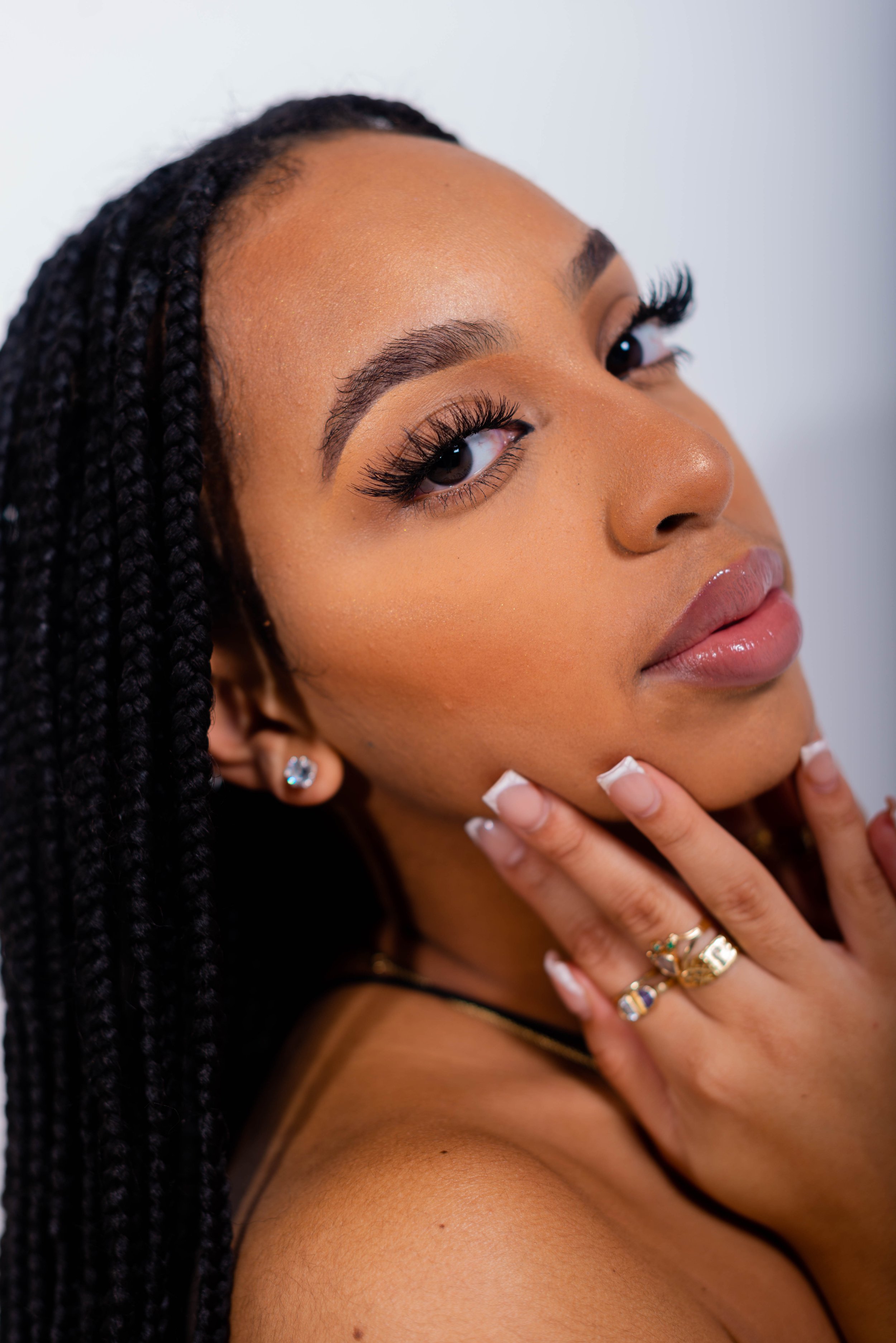 Close-up of a woman with braided hair, wearing makeup, earrings, and rings, touching her face with her hand.