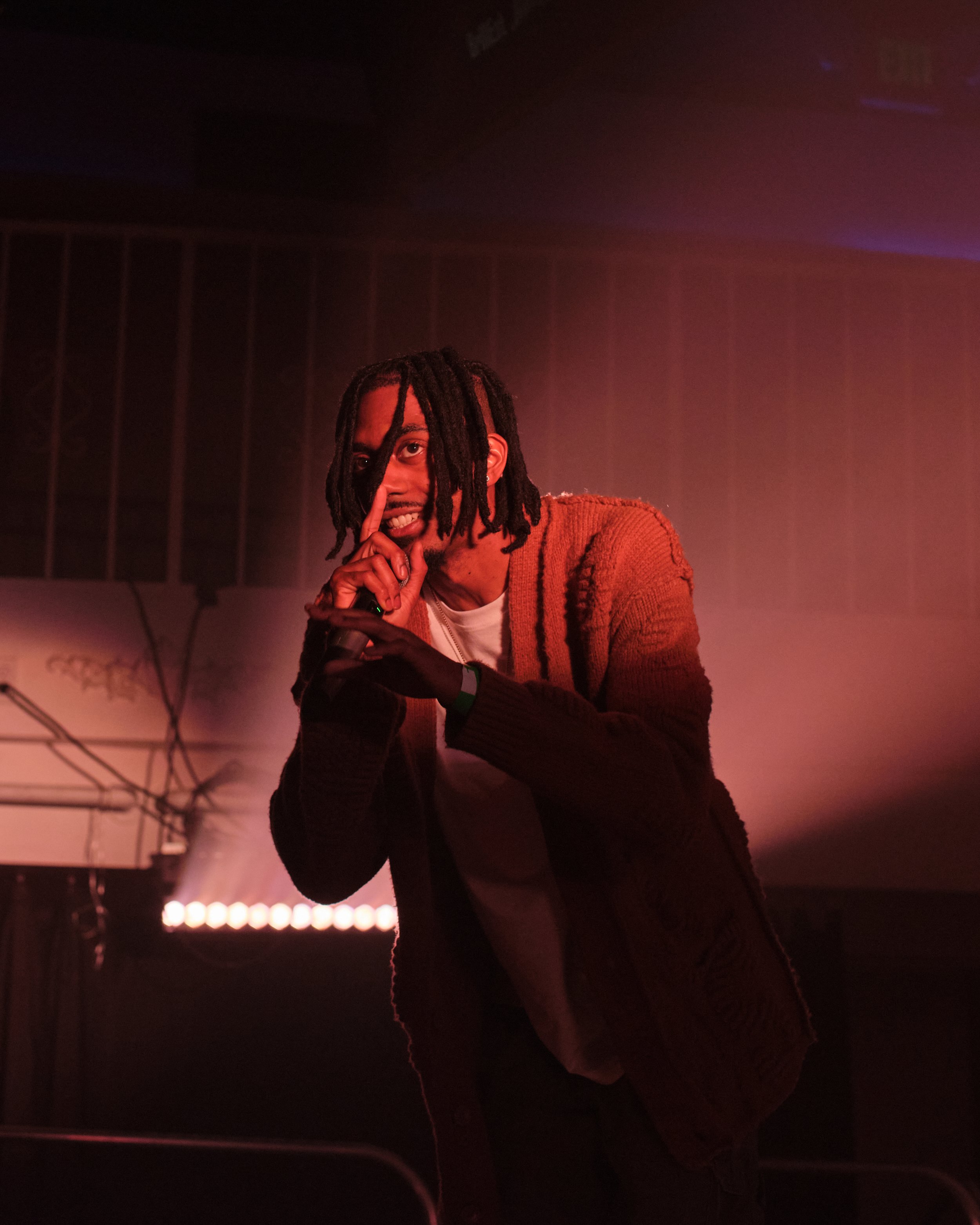 A male performer with dreadlocks on stage, holding a microphone, with one finger to his lips, in front of stage lighting and equipment.