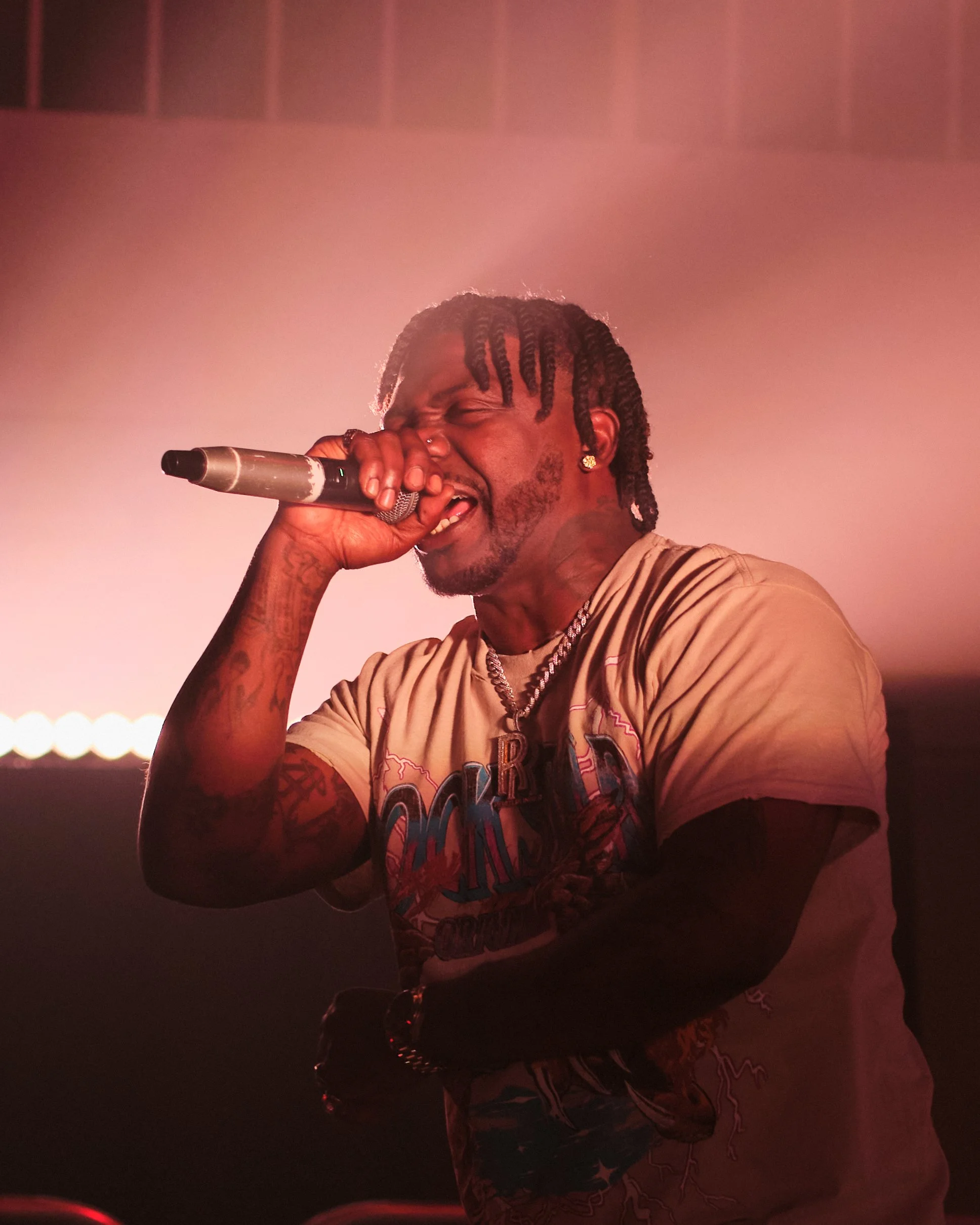 A male performer singing passionately into a microphone on stage, with closed eyes and a dynamic expression, wearing a beige graphic t-shirt, gold jewelry, and sporting braided hair.