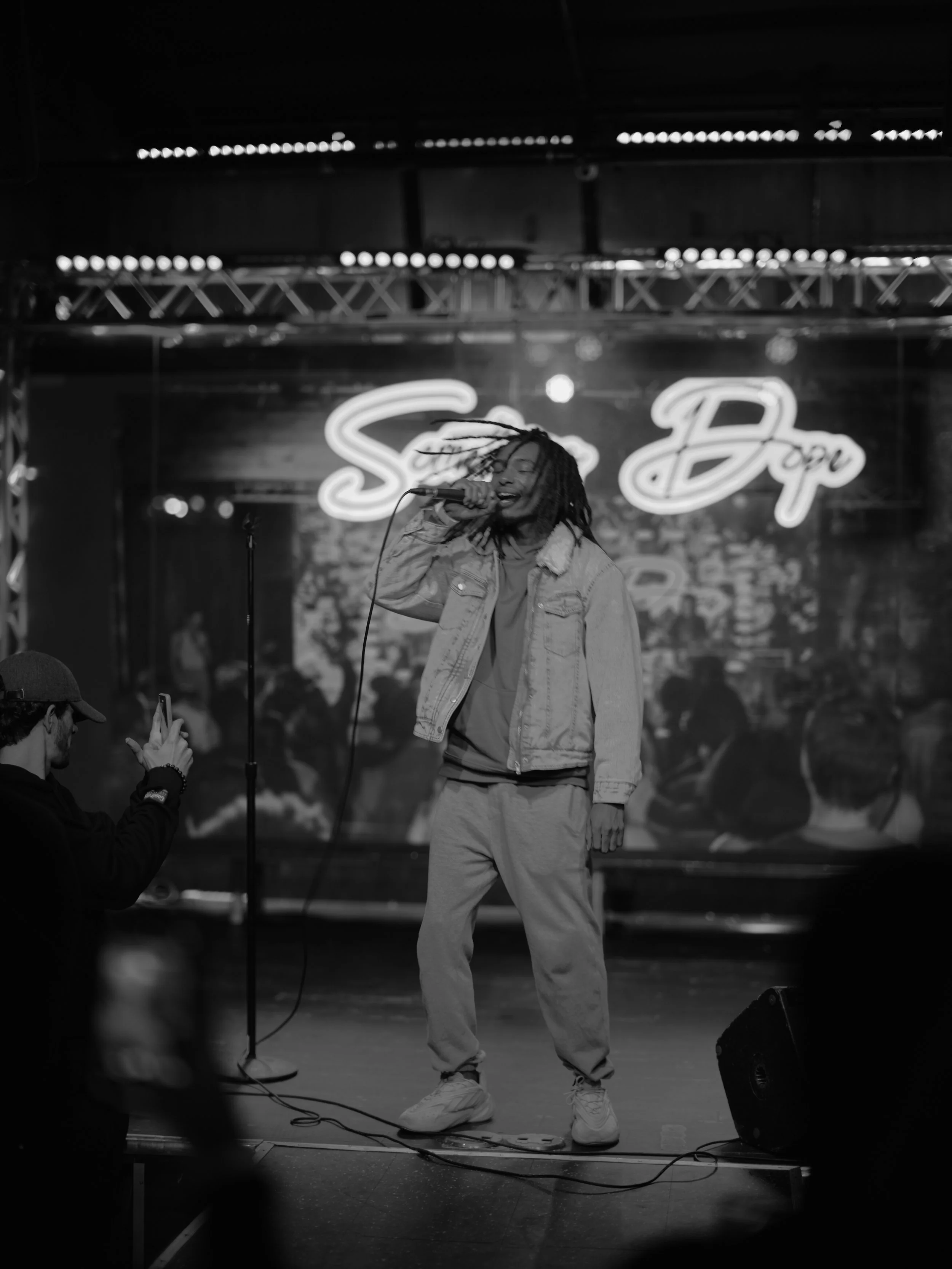 A person singing on stage with dreadlocks, wearing a denim jacket, sweatpants, and sneakers, while a man in the foreground takes a photo. The background features a neon sign with the word 'Supa' and a blurred audience.