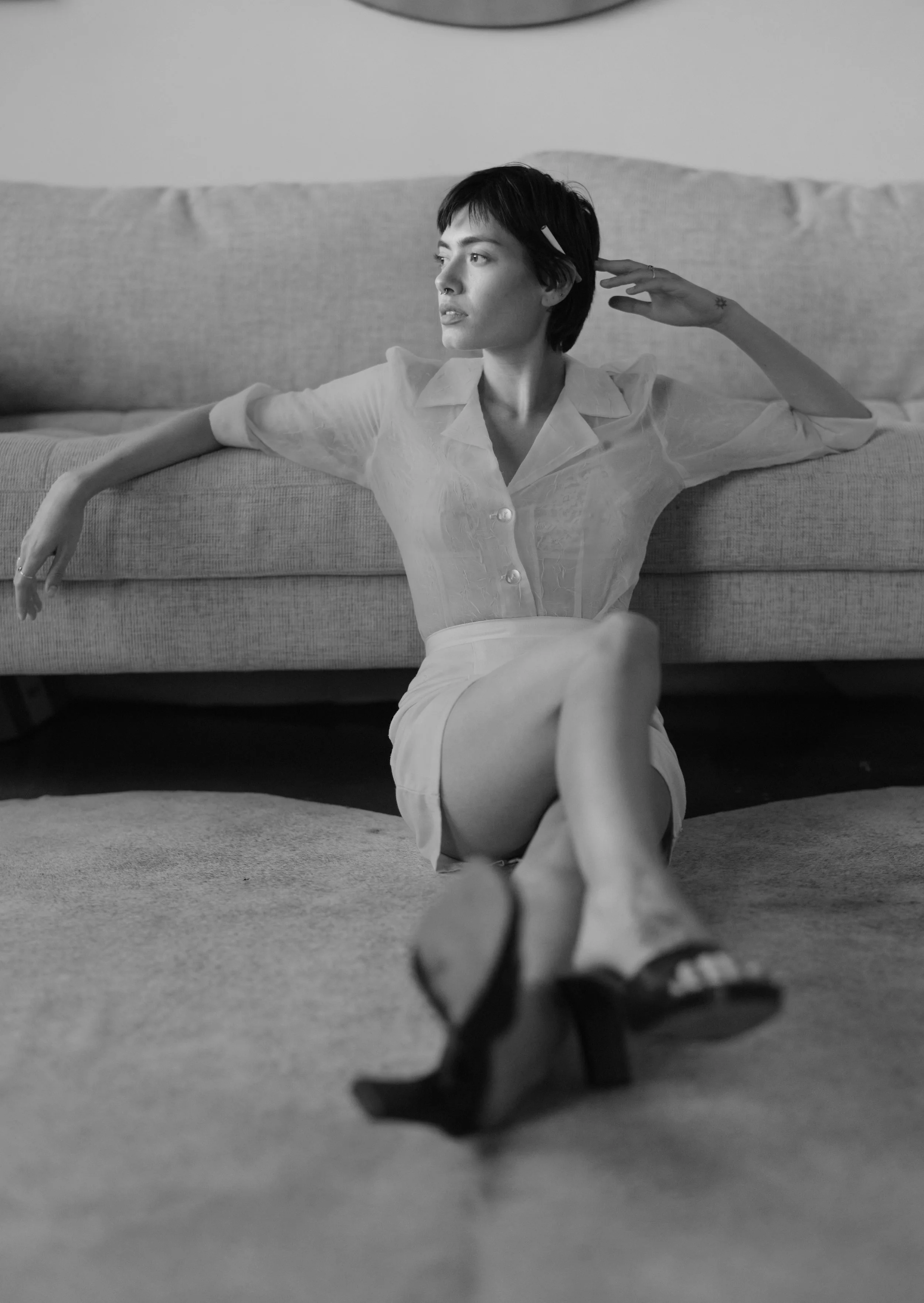 A black and white photo of a woman sitting on the floor in front of a sofa, with her legs crossed and leaning back against the sofa. She has short dark hair, is wearing a light-colored button-up shirt and high-heeled shoes, and is looking to her left