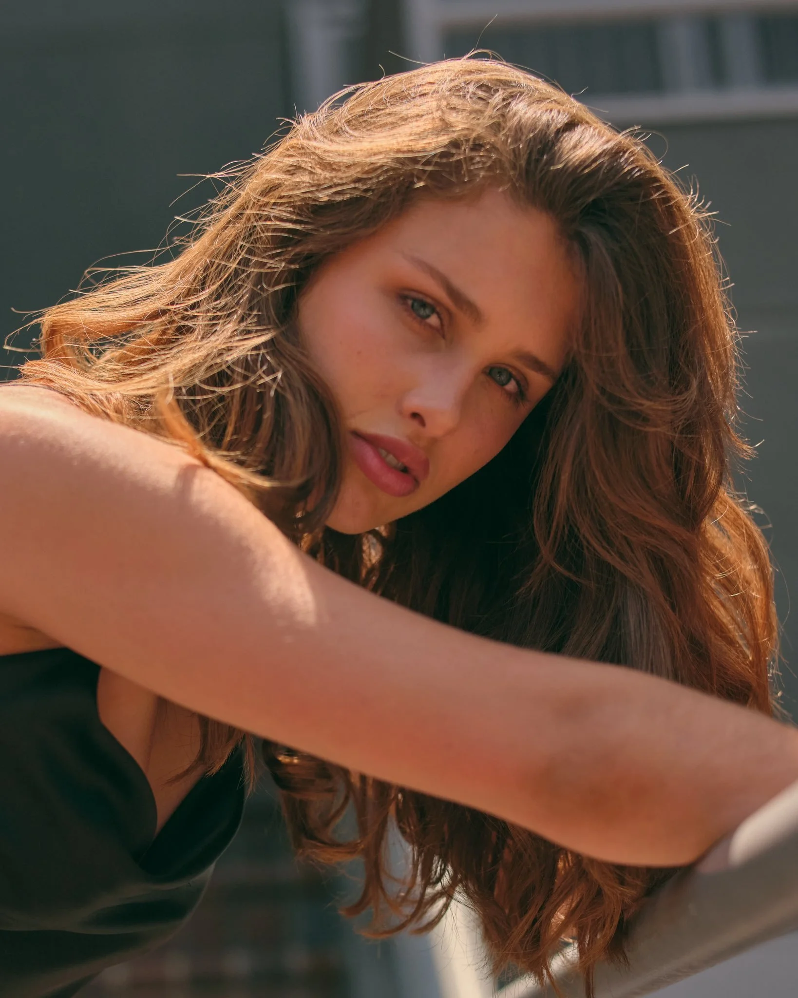 Close-up of a woman with long brown wavy hair, looking at the camera with a serious expression, outdoors in sunlight.