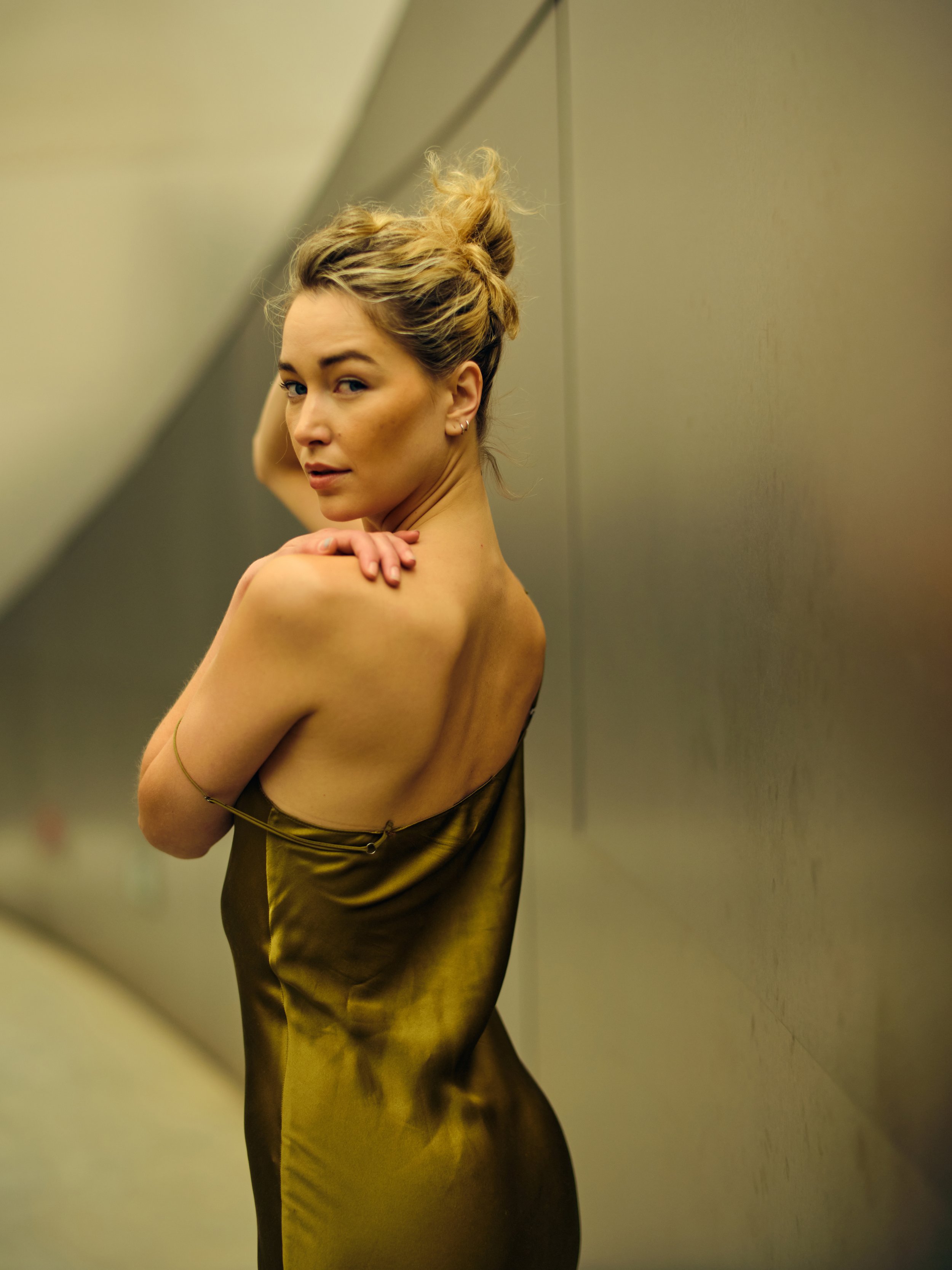 A woman in a satin gold dress standing against a metallic wall, looking over her shoulder with styled hair.
