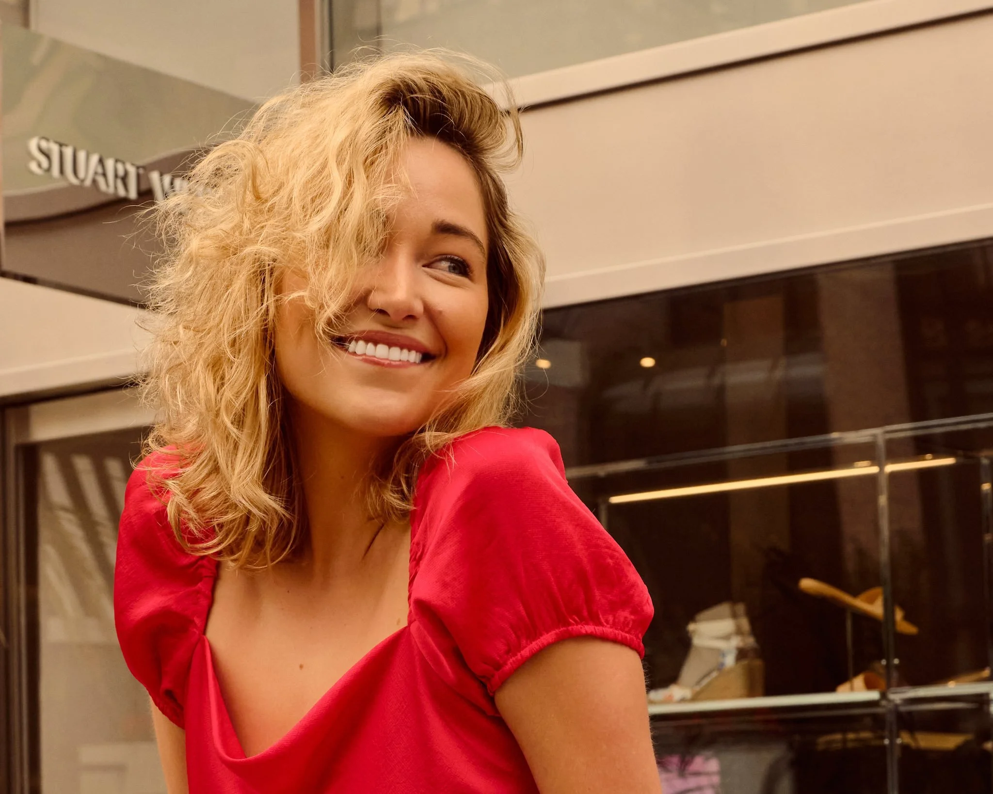 A young woman with shoulder-length curly blonde hair, smiling and looking to her right, wearing a red top, standing outdoors near a storefront.
