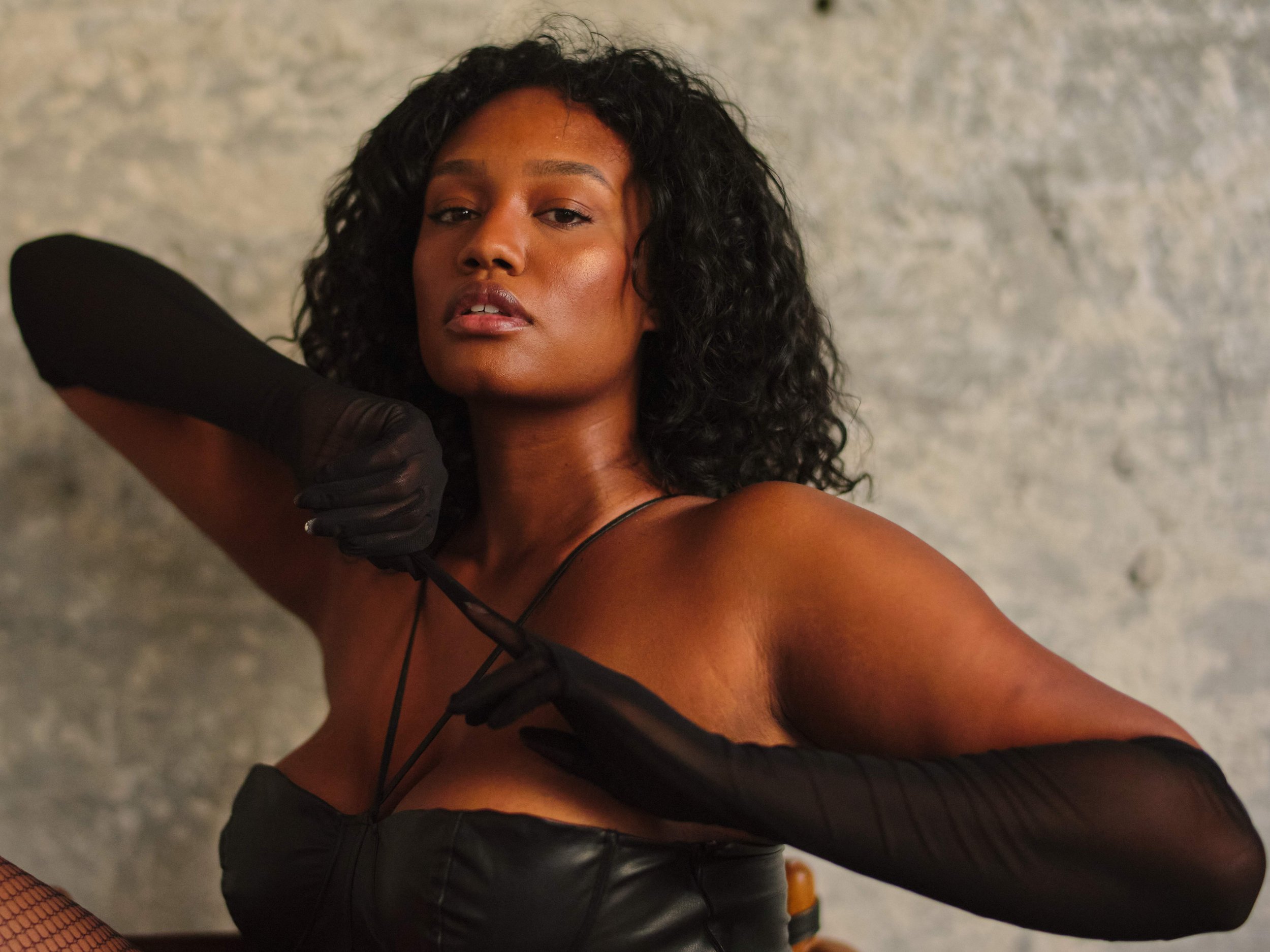 A woman with curly dark hair posing confidently in a black leather top and black gloves against a textured gray wall.