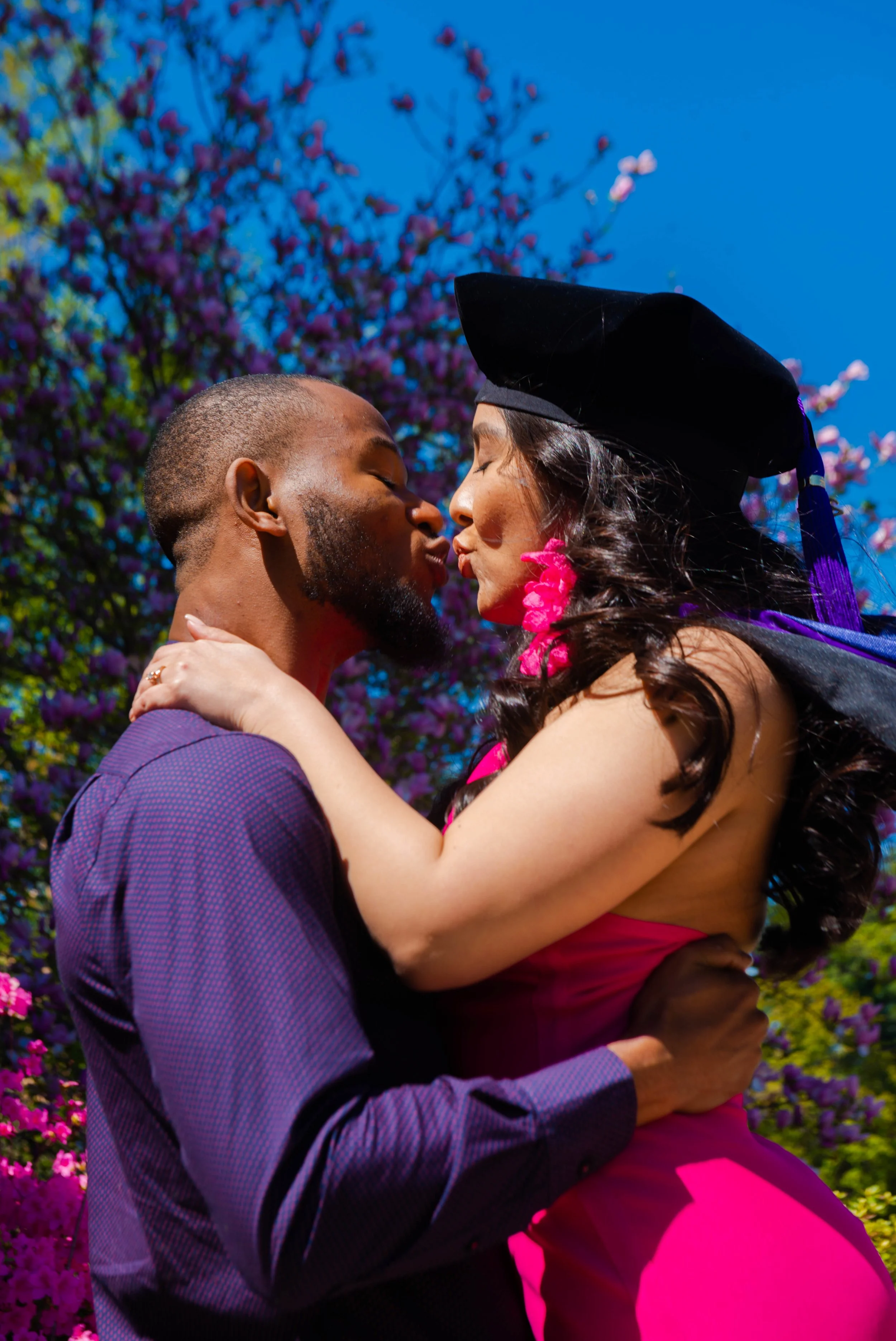 A couple is embracing and kissing outdoors on a bright, sunny day, with purple flowering trees and blue sky in the background. The woman is wearing a graduation cap and gown, and the man is holding her as they kiss.