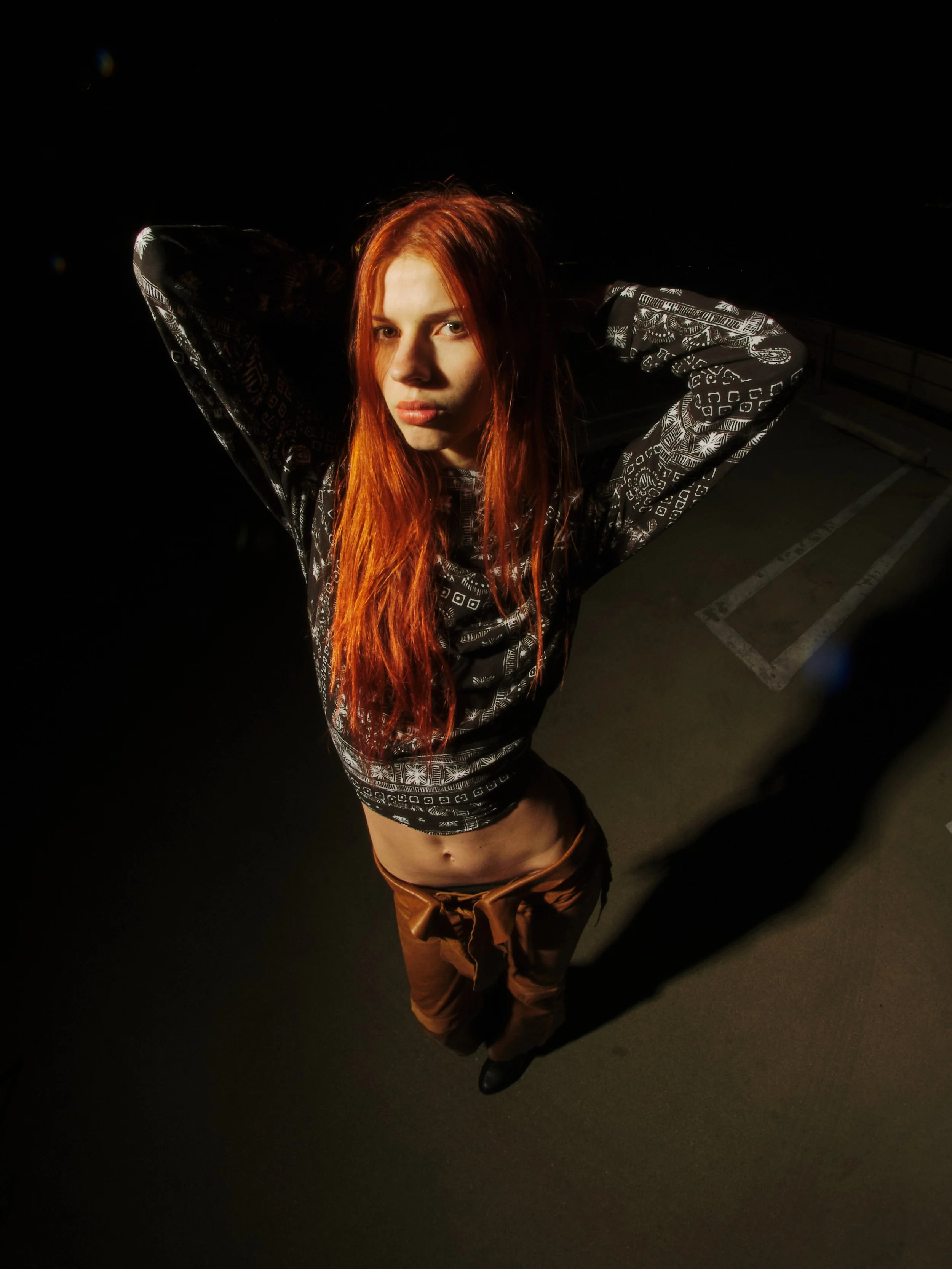 Young woman with long red hair standing in a dark parking lot, with her arms raised behind her head, illuminated by a single light source.