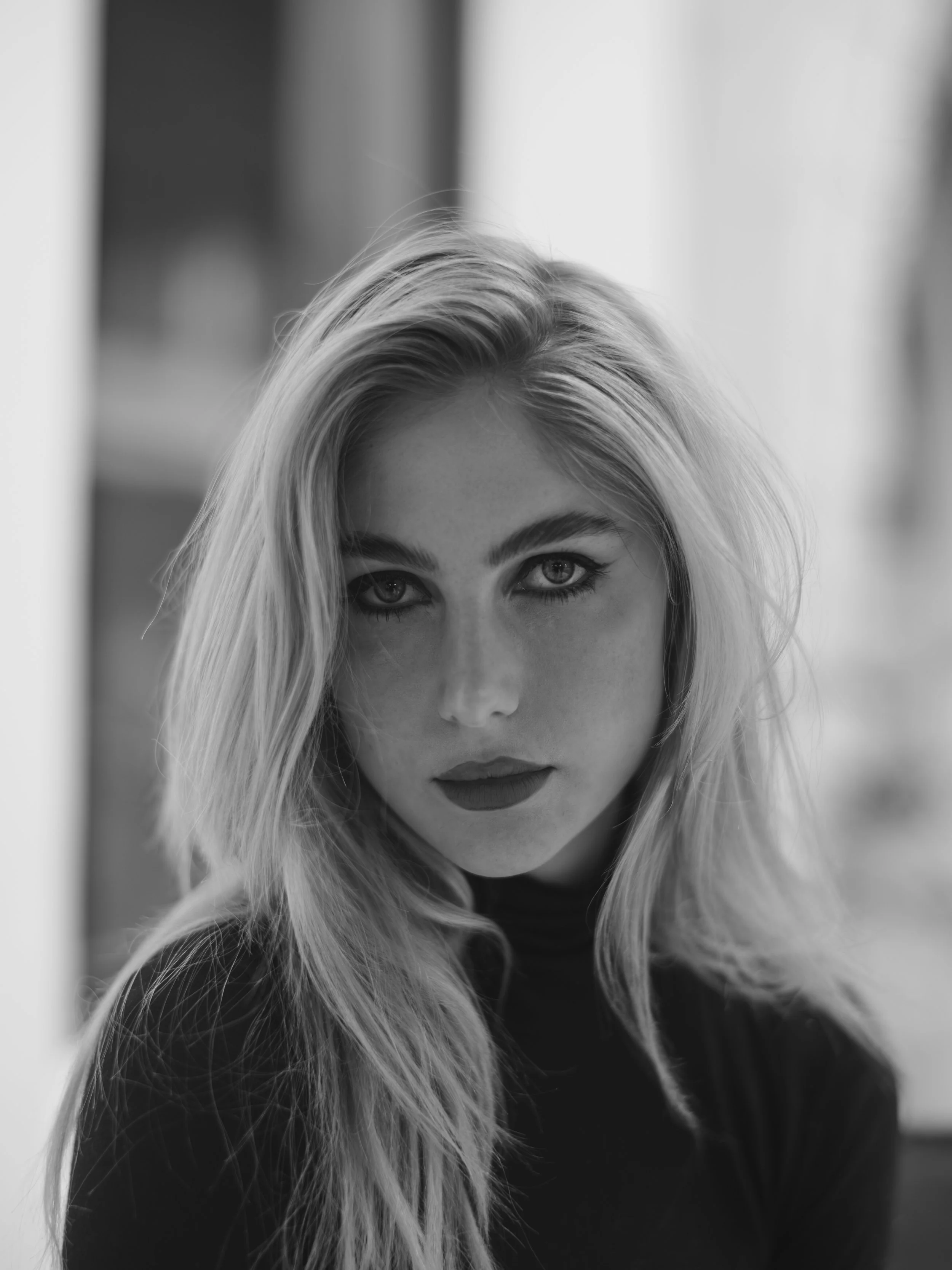 Black-and-white photo of a young woman with wavy blonde hair, wearing a dark top, looking directly at the camera with a serious expression.
