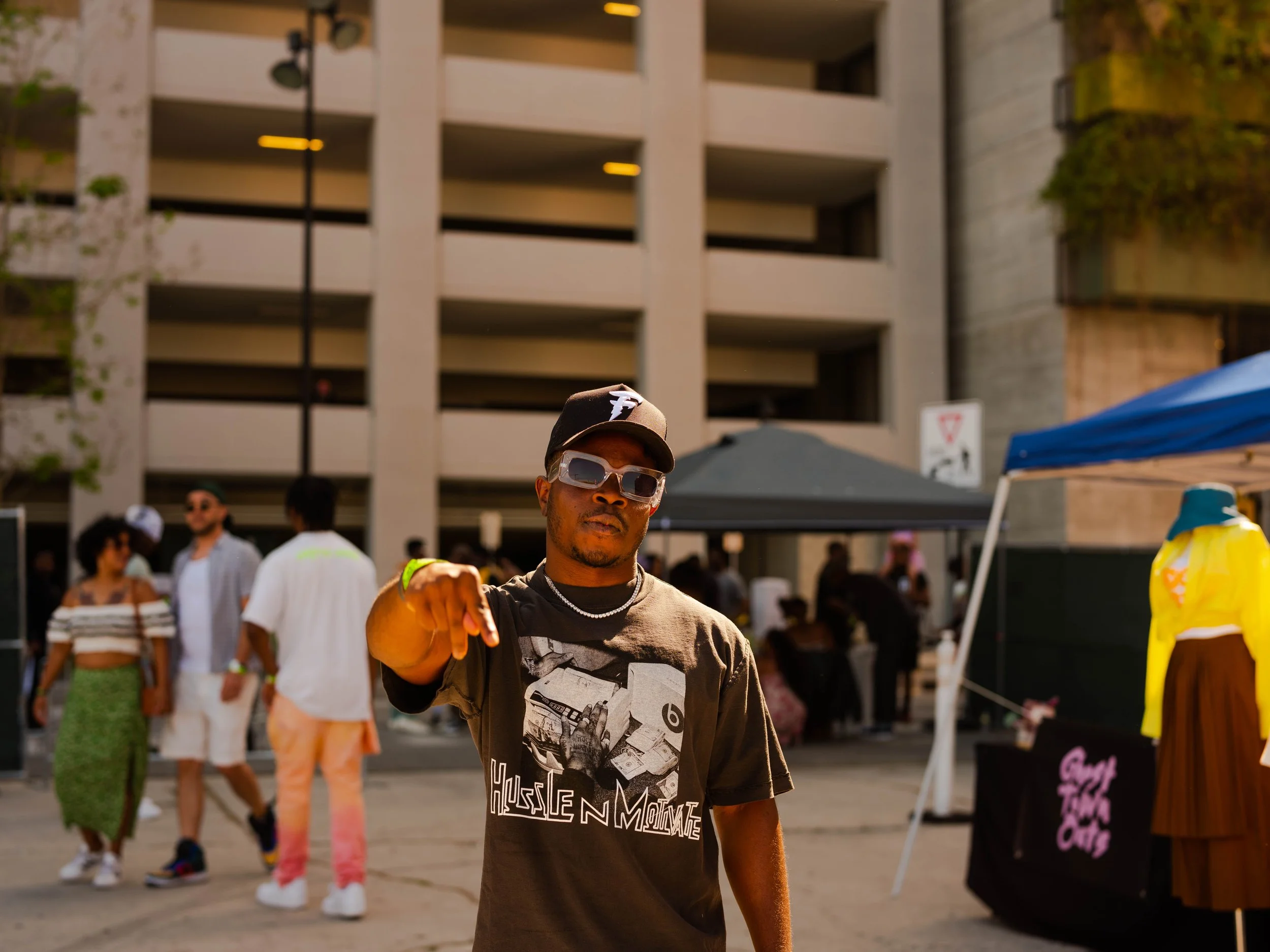 A man wearing sunglasses, a black baseball cap, and a graphic t-shirt pointing towards the camera at an outdoor event, with people, tents, and buildings in the background.