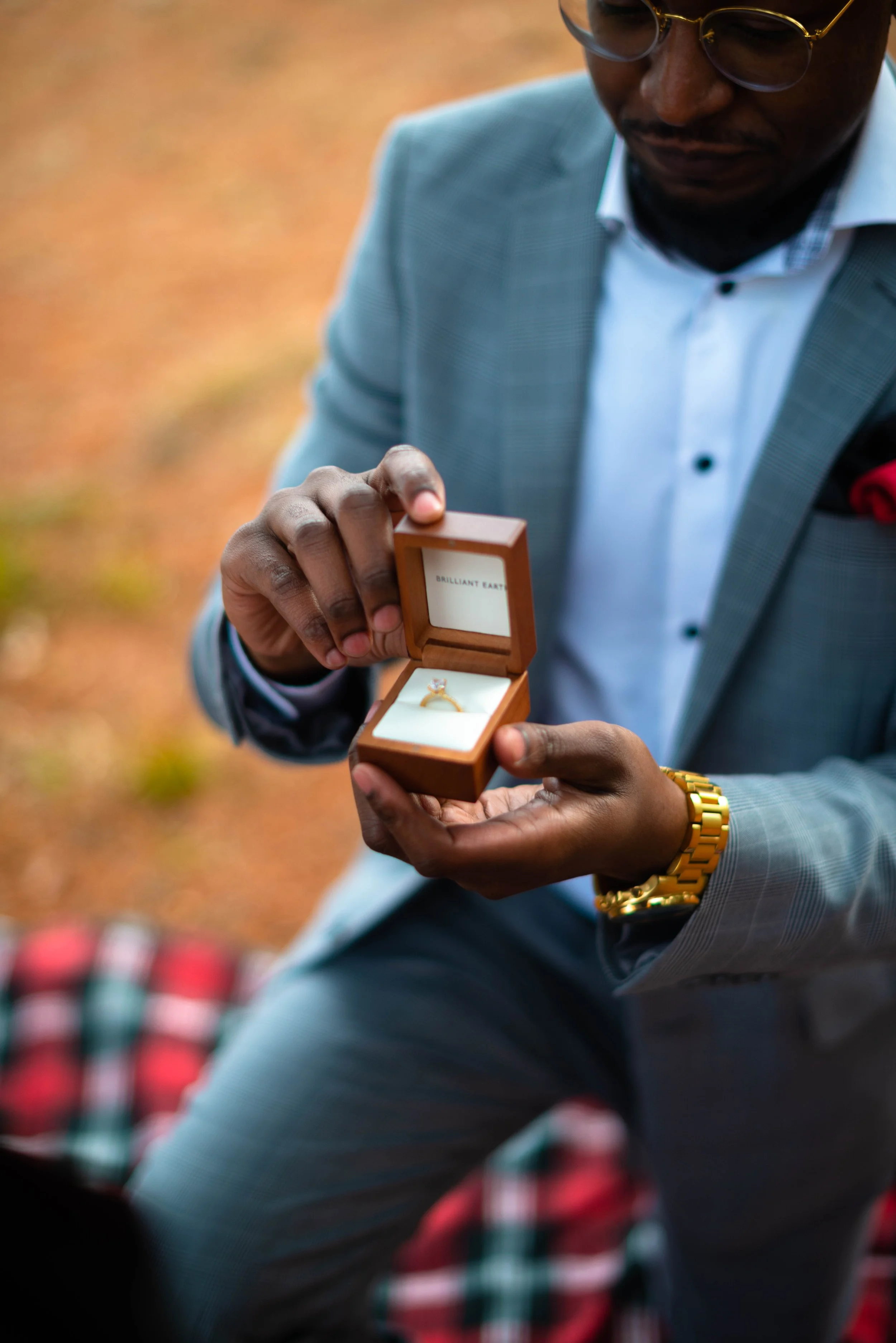 A man in a gray suit holding a small wooden jewelry box with a ring inside, outside on a fall day.