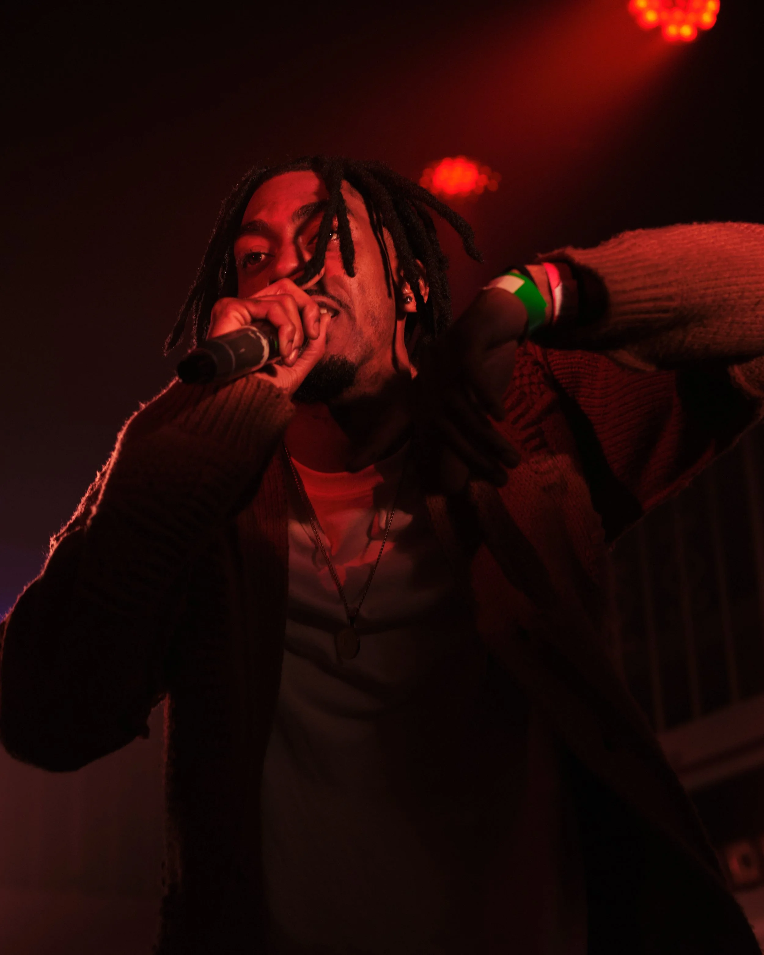 A male performer with dreadlocks singing into a microphone on stage with red lighting.