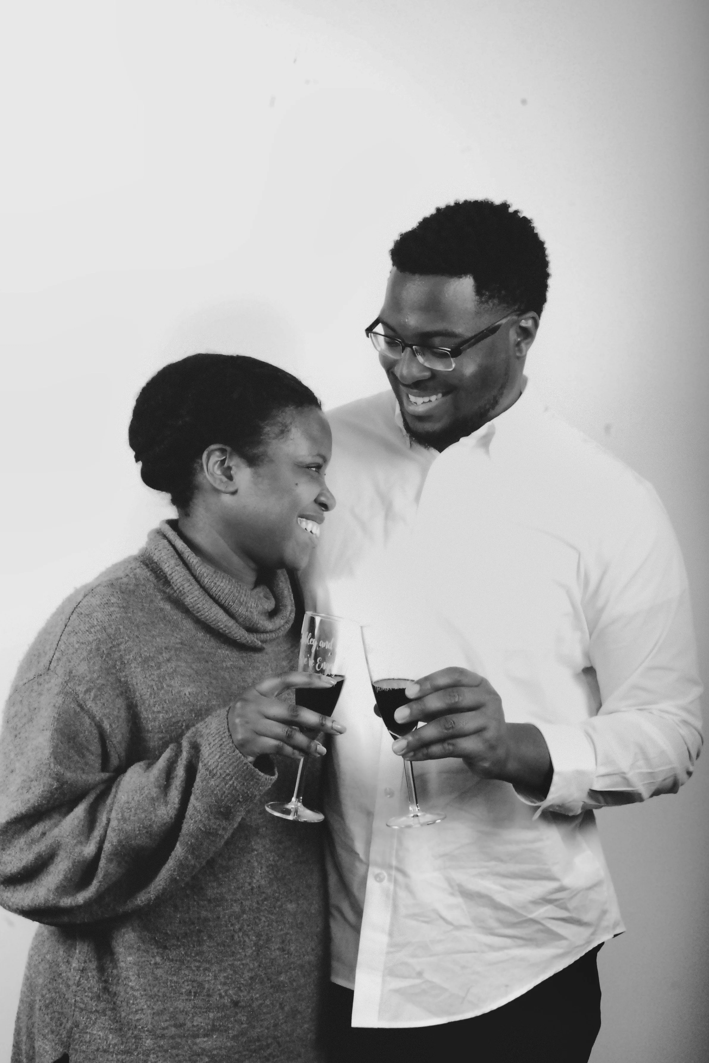 A happy couple smiling, holding glasses of red wine, celebrating together, with the woman wearing a turtleneck sweater and the man in a white shirt, standing against a plain background.