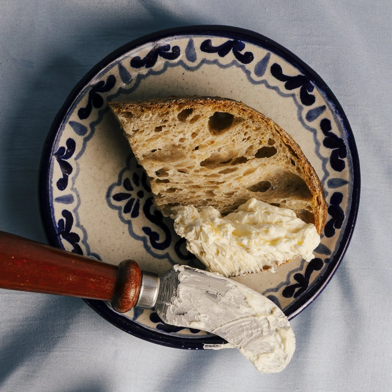 A ceramic bowl with blue decorative patterns containing a slice of rustic bread and a serving of cream cheese, with a butter knife resting on the edge of the bowl.