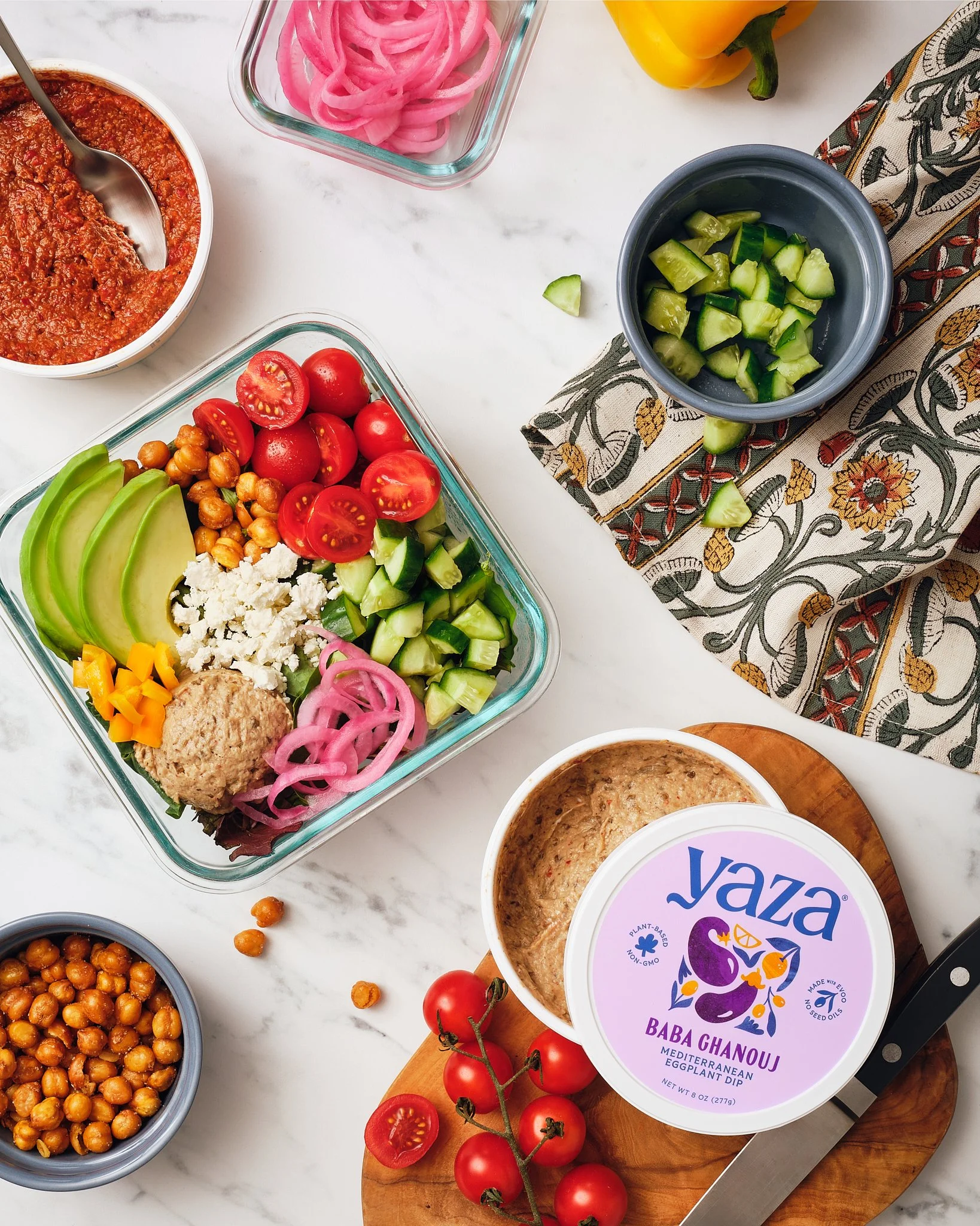 An assortment of fresh vegetables, hummus, and chickpeas on a white countertop, ready to make a Mediterranean-inspired meal.