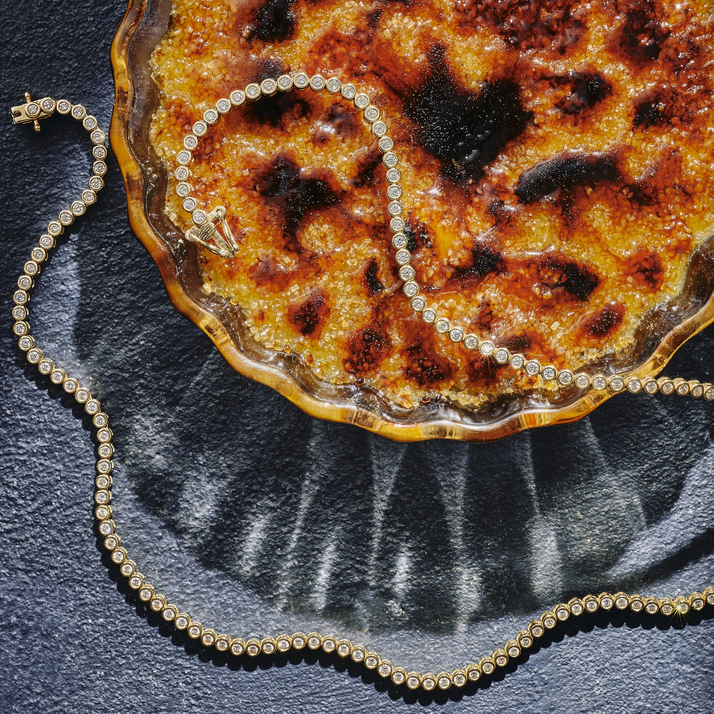 A round, textured, amber-colored dish with charred spots, garnished with a sparkly diamond necklace and a matching bracelet, all set on a dark, textured surface.