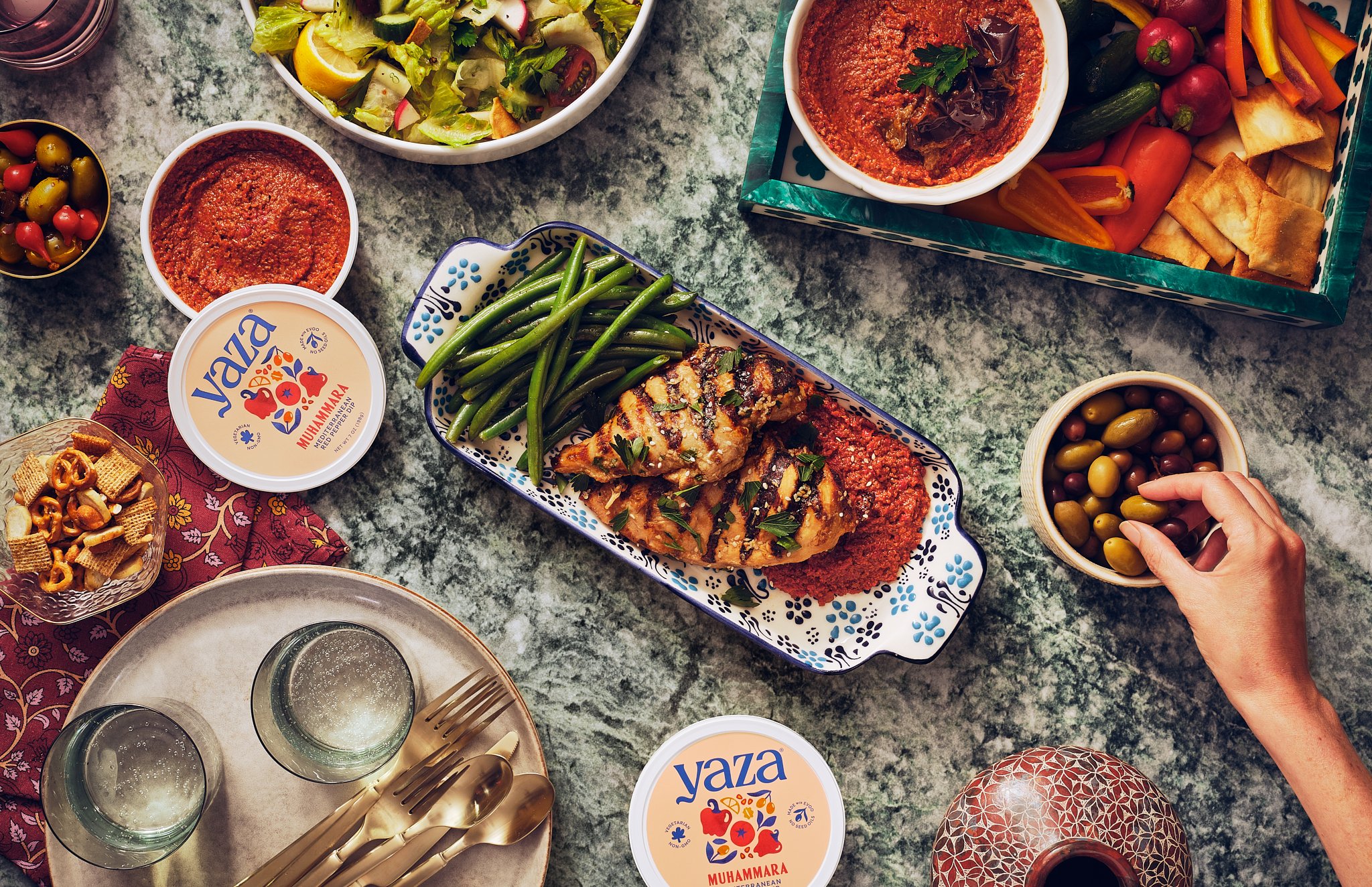 A table of Mediterranean food including bowls of hummus, salads, olives, grilled fish with green beans, and various vegetable snacks. There are also plates with utensils, a scented oil diffuser, and boxed snacks.