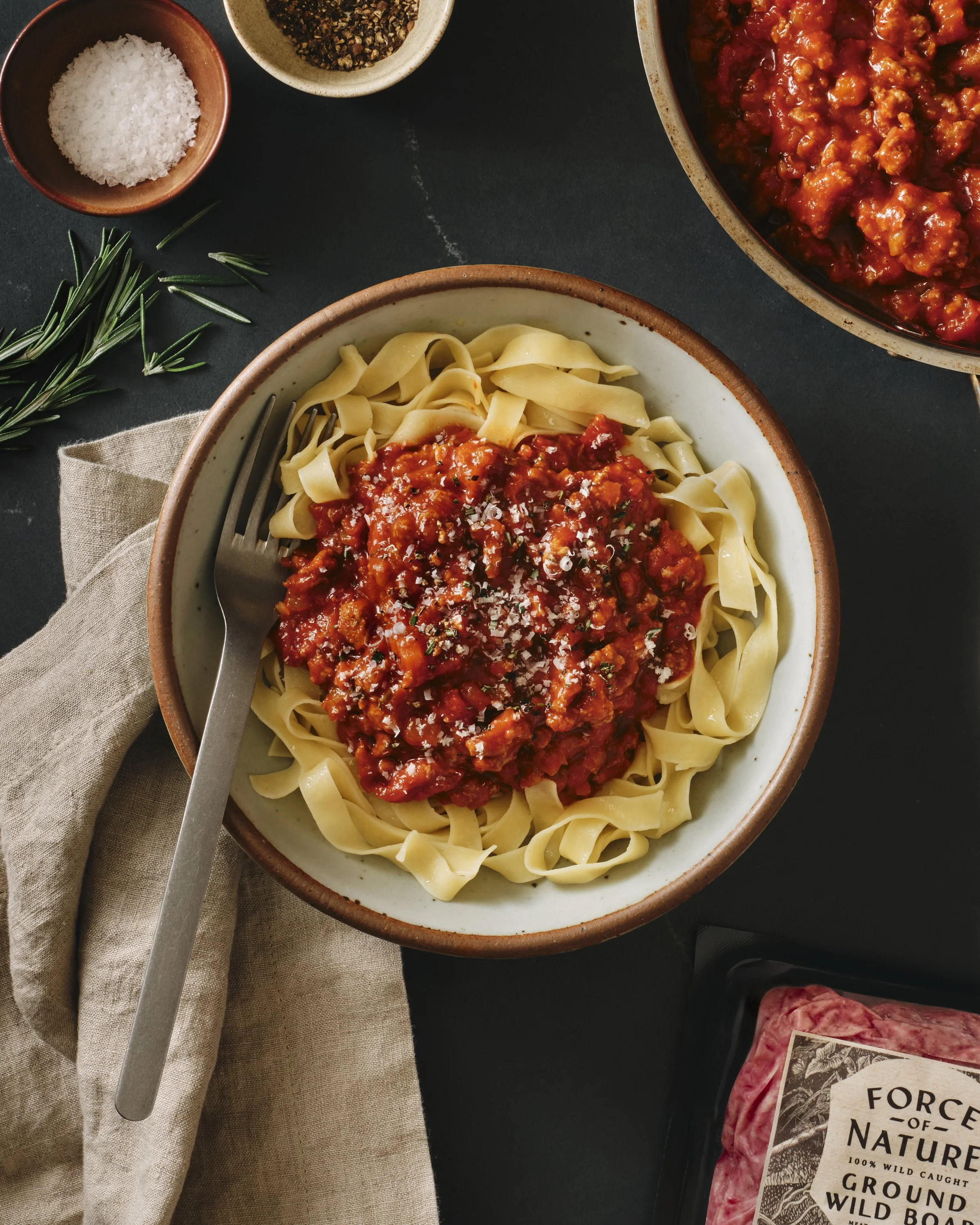 A bowl of cooked pasta with tomato sauce, garnished with grated Parmesan cheese, on a dark table with a fork, a sprig of rosemary, and small bowls of salt and pepper nearby.