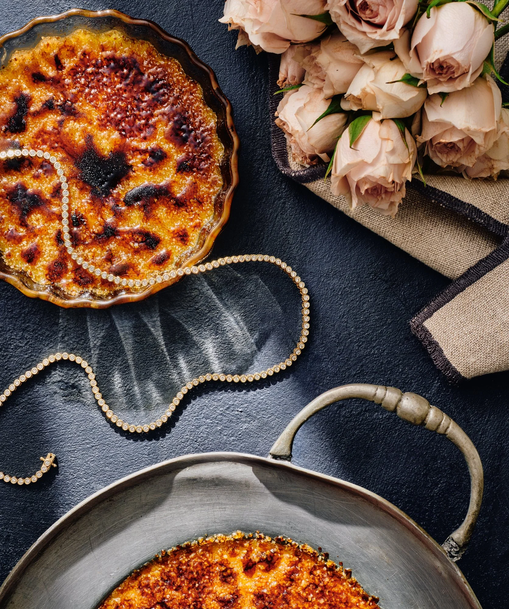 A baked dish in a fluted ceramic dish, next to a bouquet of pale pink roses and a pearl necklace, on a black surface.