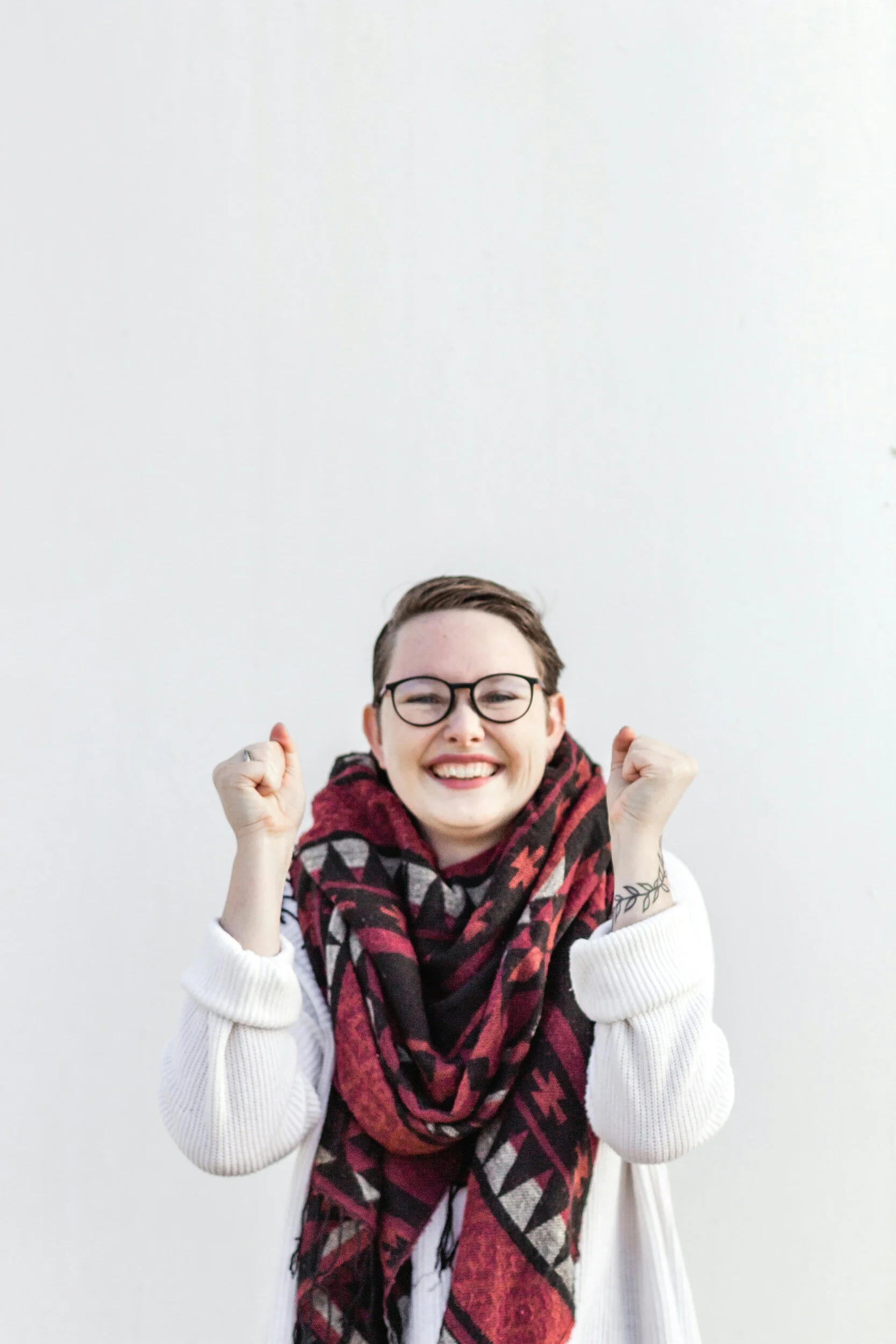 A person with short hair, glasses, white sweater, and a red, black, and white patterned scarf smiling and celebrating with fists raised in front of a plain white wall.