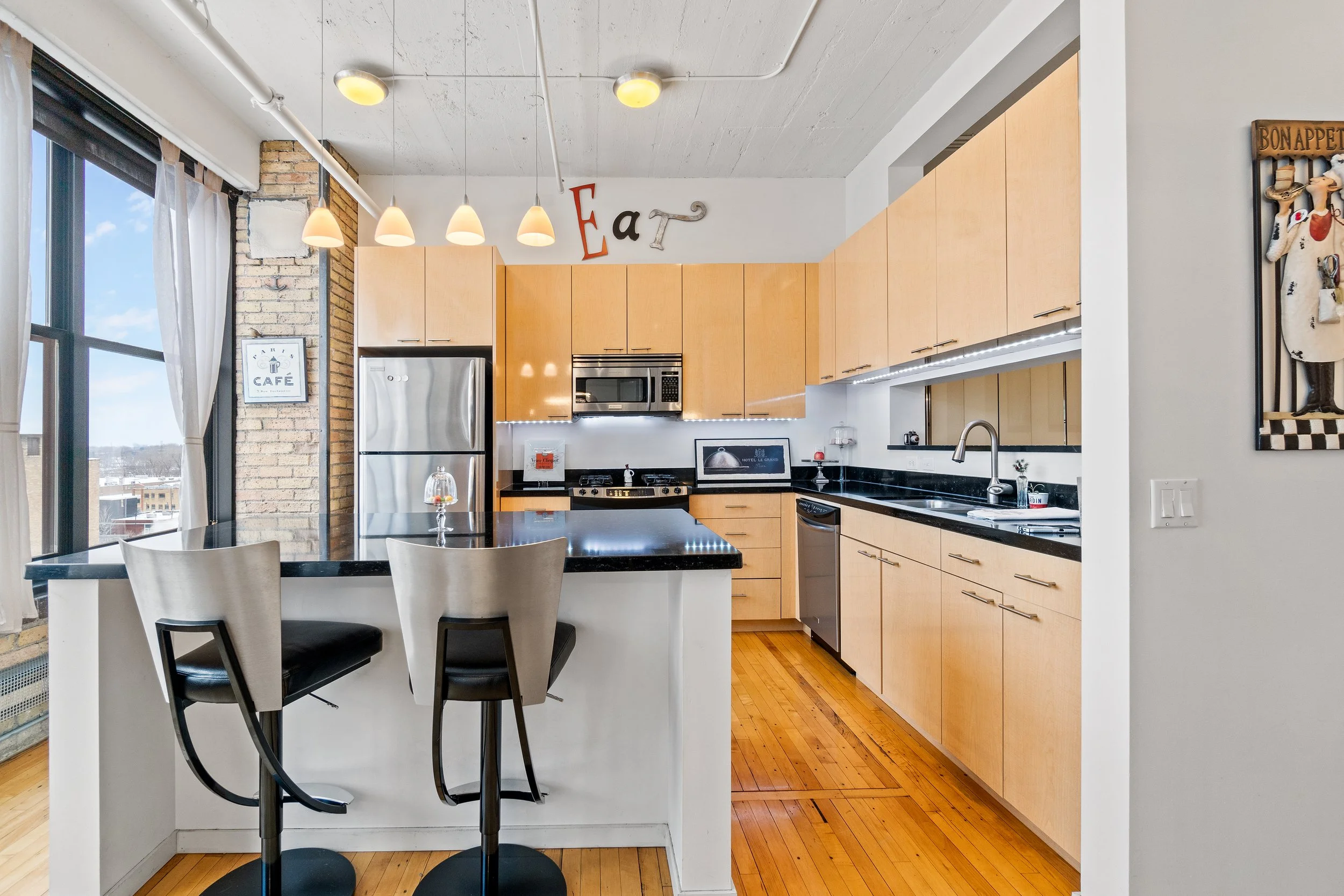 Modern kitchen with light wood cabinets, black countertops, stainless steel appliances, and a breakfast bar with two tall chairs. Large window with white curtains, exposed brick wall.