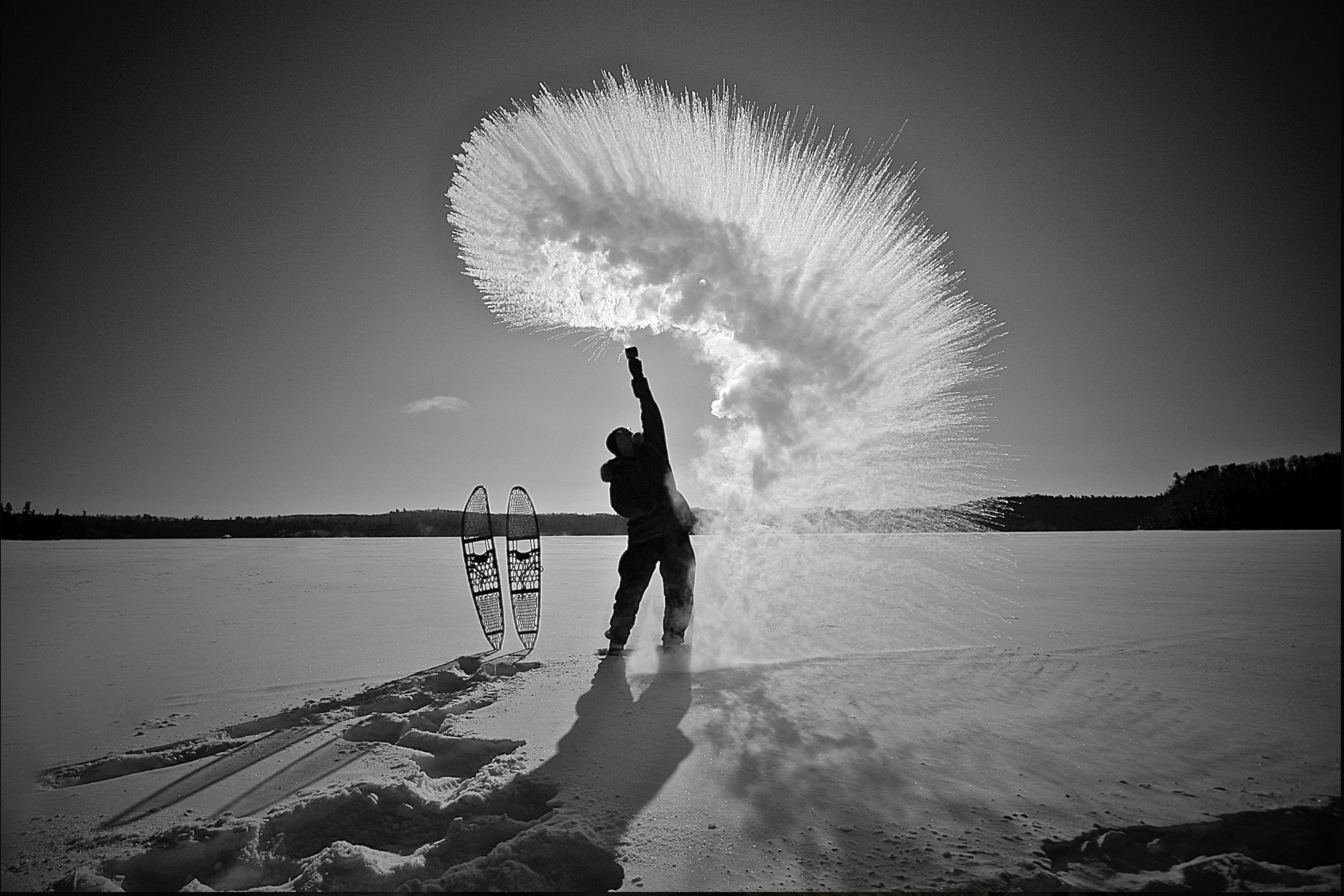 A person standing on snow-covered ground, creating an explosion of snow in an arc, with two snowshoes lying nearby, in a vast, open, snow-covered landscape under a clear sky.