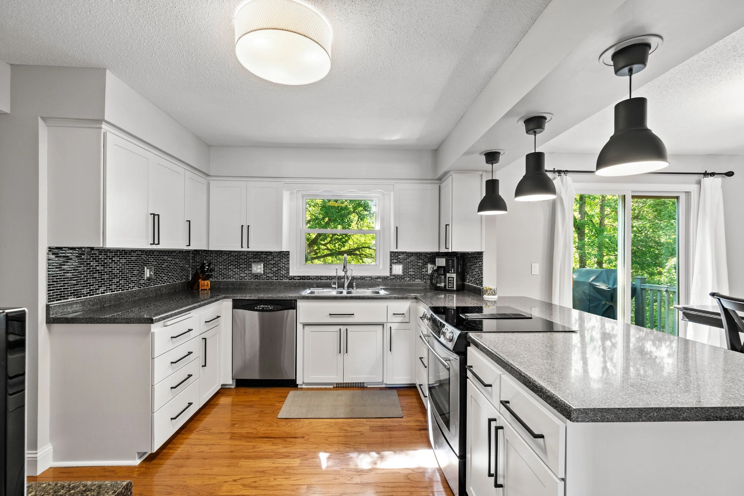 Modern kitchen with white cabinets, black backsplash, gray countertops, and black pendant lights, overlooking a green outdoor area through sliding glass doors.