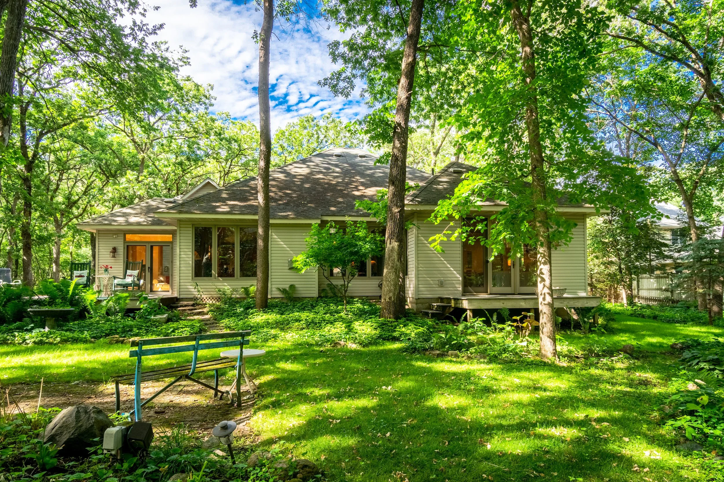 A house with a deck surrounded by lush green trees and a well-maintained garden and lawn in a sunny backyard.