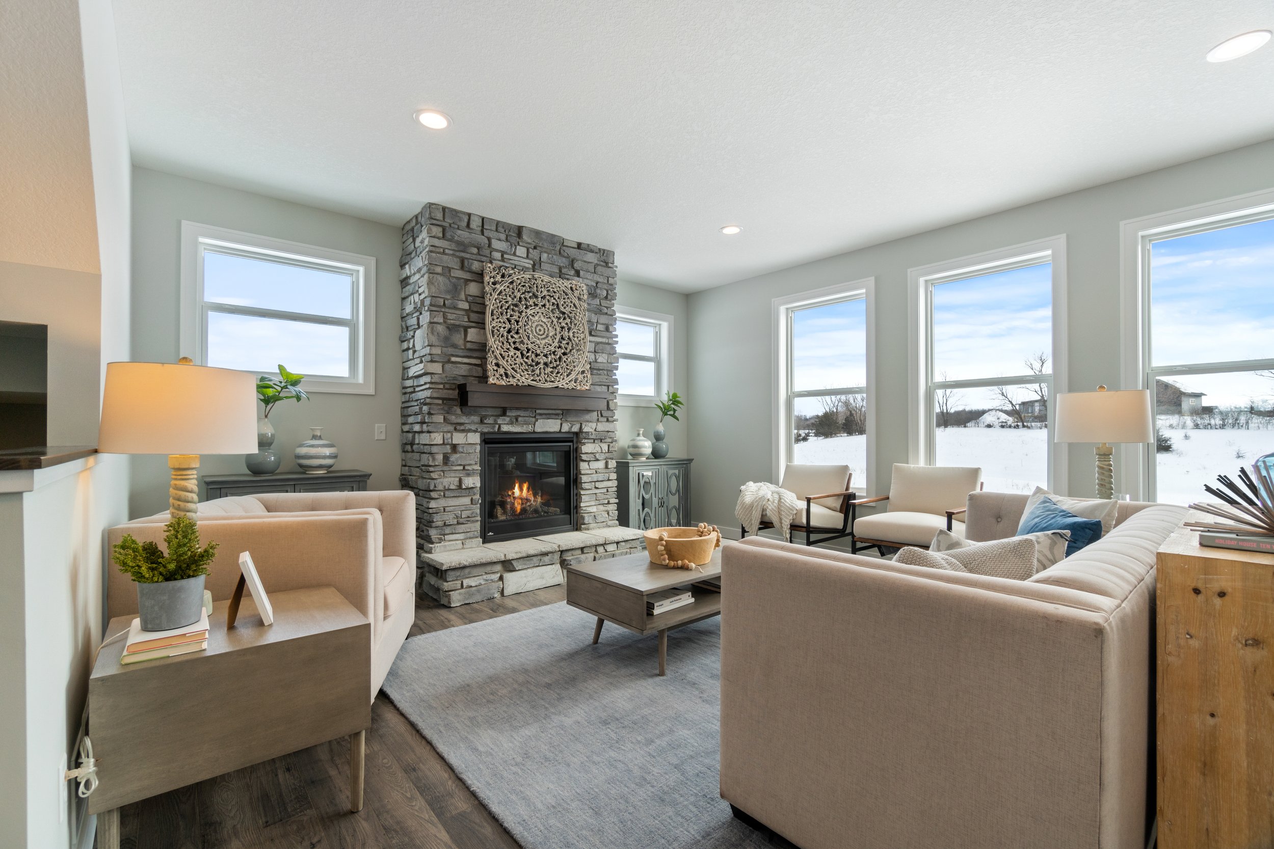 Living room with a stone fireplace, beige sofas, wooden coffee table, large windows showing snow outside, and a cozy winter scene.