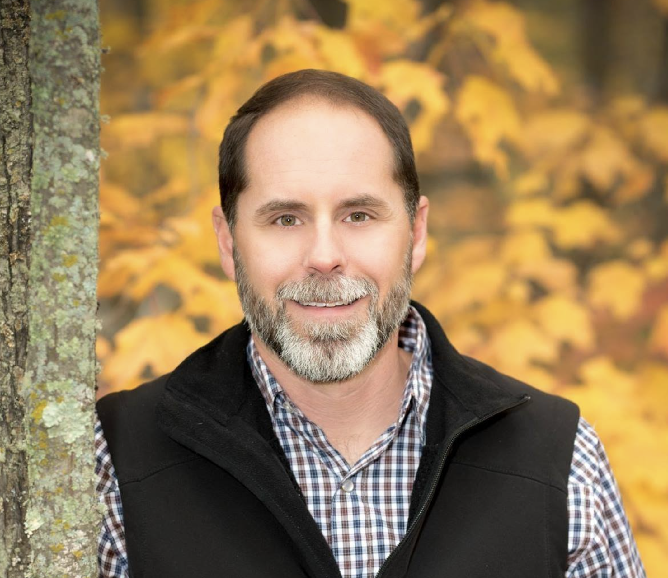 A man with a beard and gray hair smiling outdoors with fall foliage in the background.