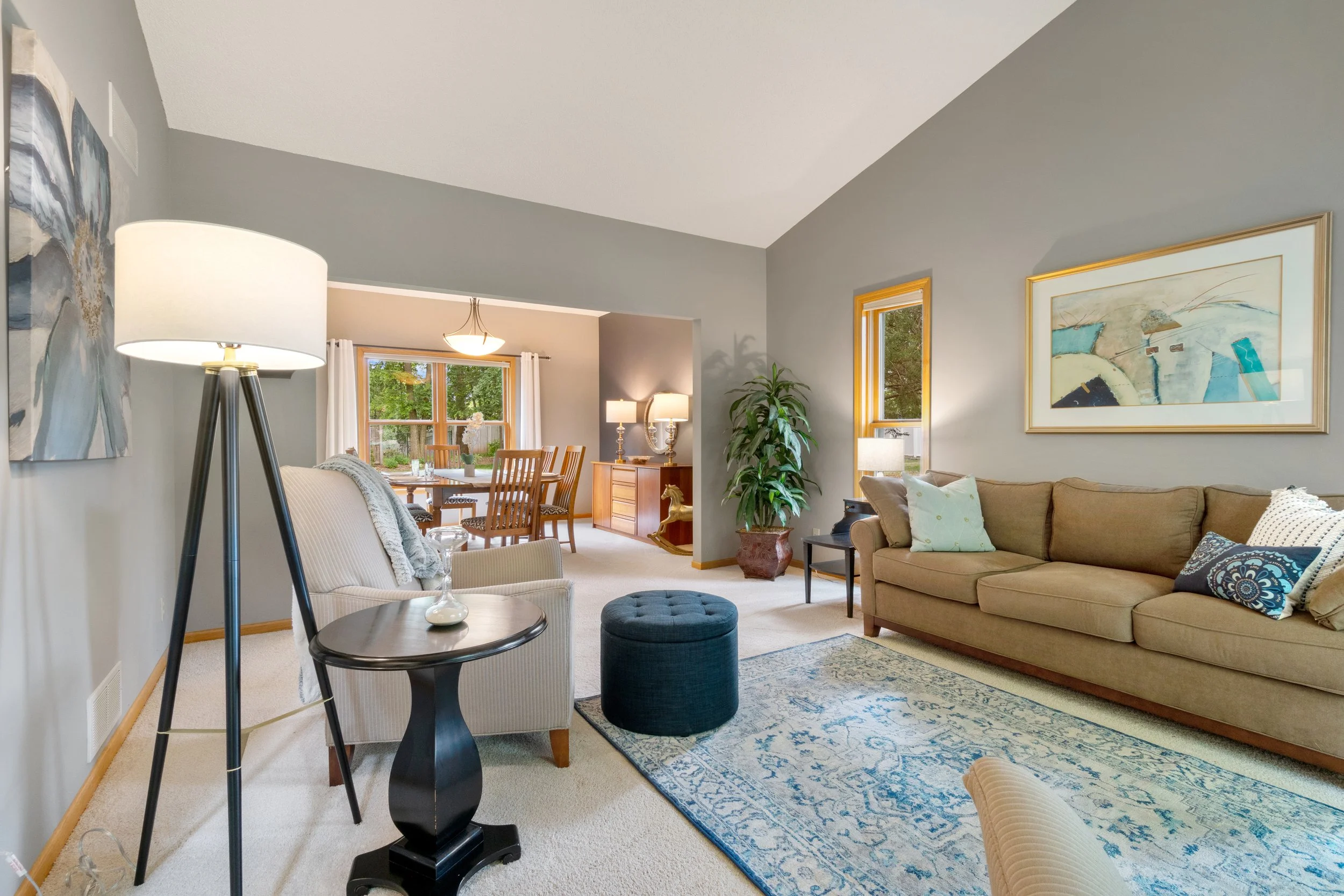 Living room with beige sofa, armchair, blue ottoman, floor lamp, large framed artwork, and a window with white curtains, leading to a dining area with a wooden table and chairs, and a sideboard with lamps.