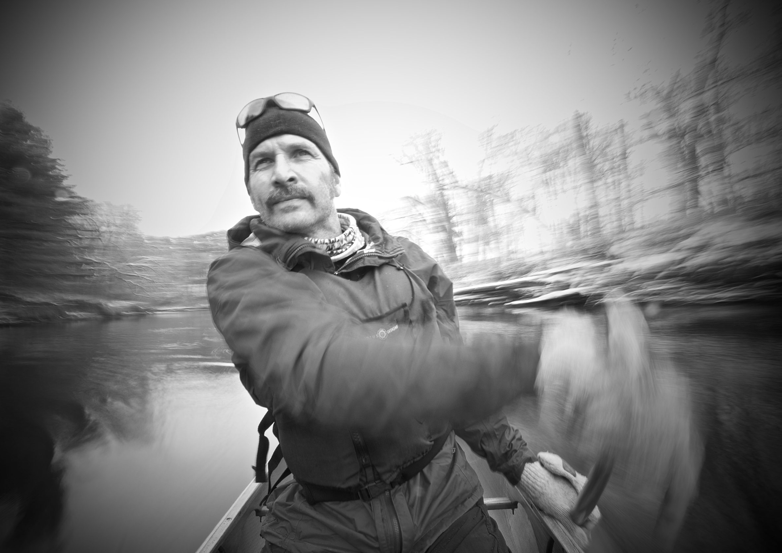 A man wearing a beanie, glasses on his head, and outdoor gear, is sitting in a boat on a river holding a fish, with trees in the background. The image is in black and white.
