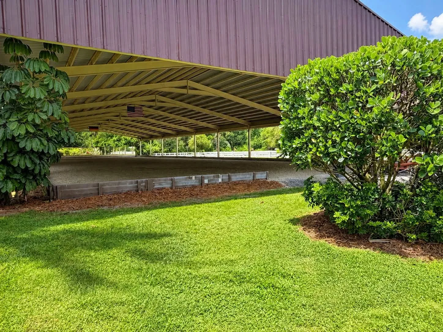 Outdoor covered arena with a dirt floor, surrounded by green grass, bushes, and trees, with a purple roof and American flag hanging from the ceiling.
