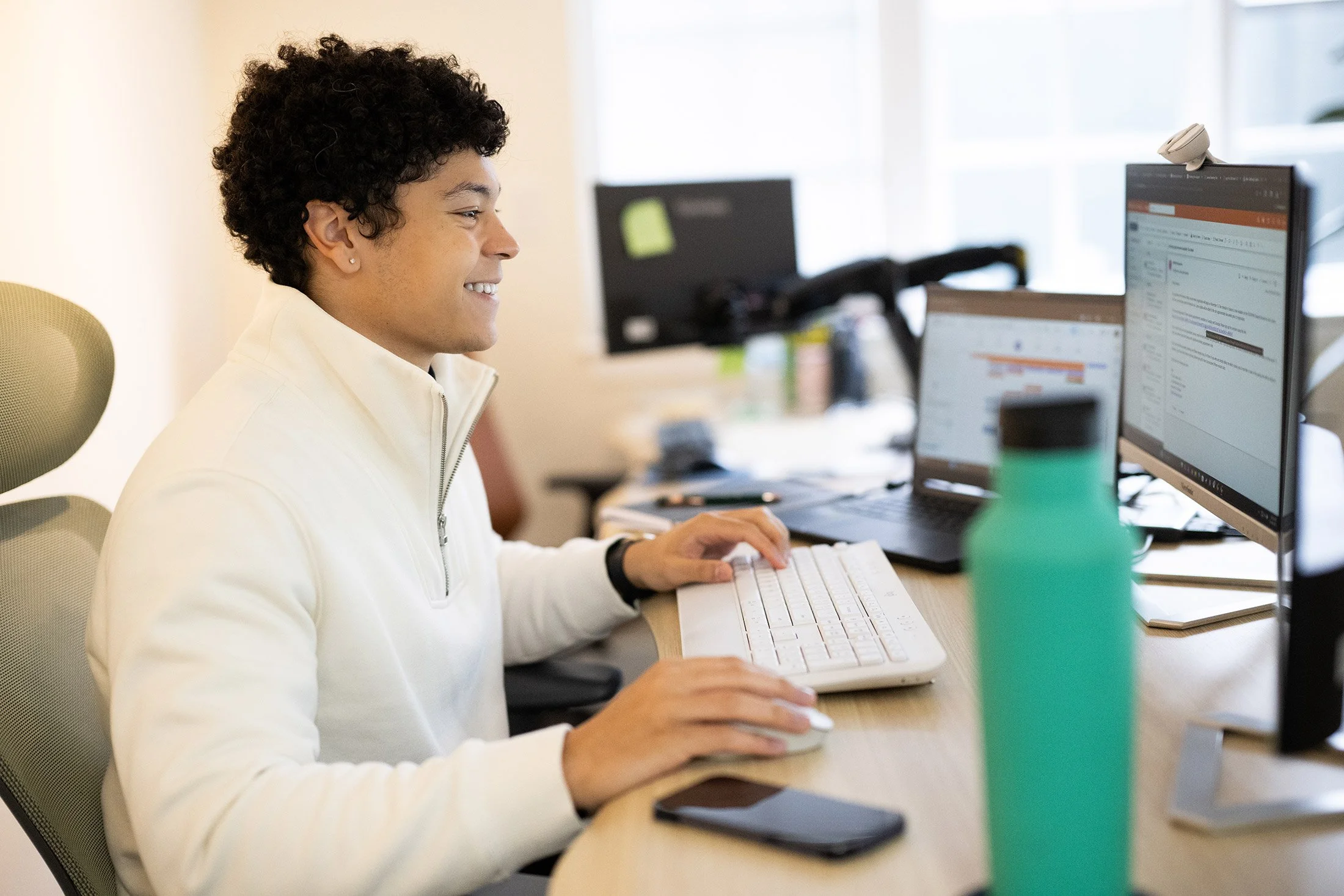 A person sitting at a desk working on a computer, smiling, with multiple screens and a water bottle on the desk.