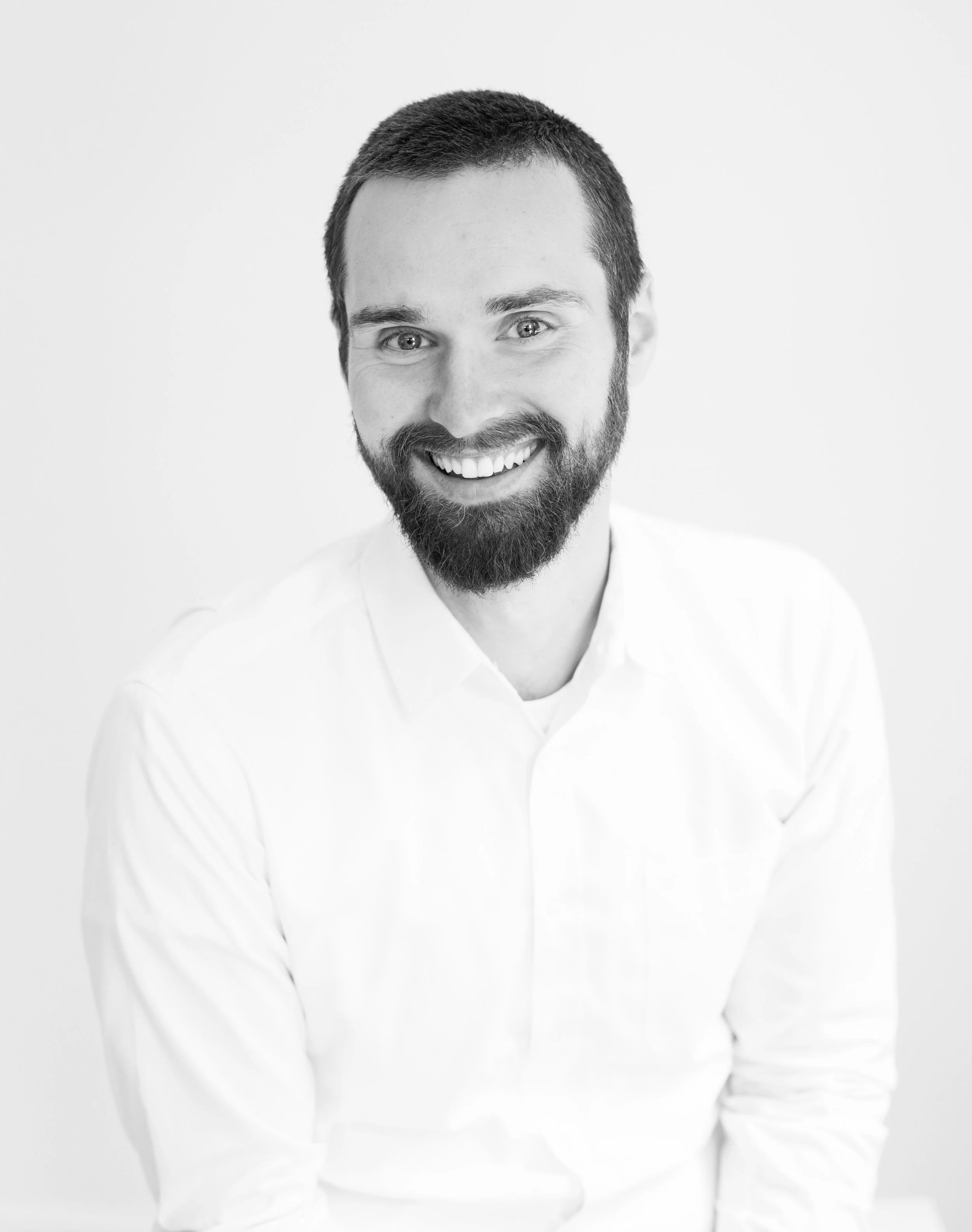 Black and white portrait of a smiling man with beard and short hair, wearing a white shirt, against a plain light background.