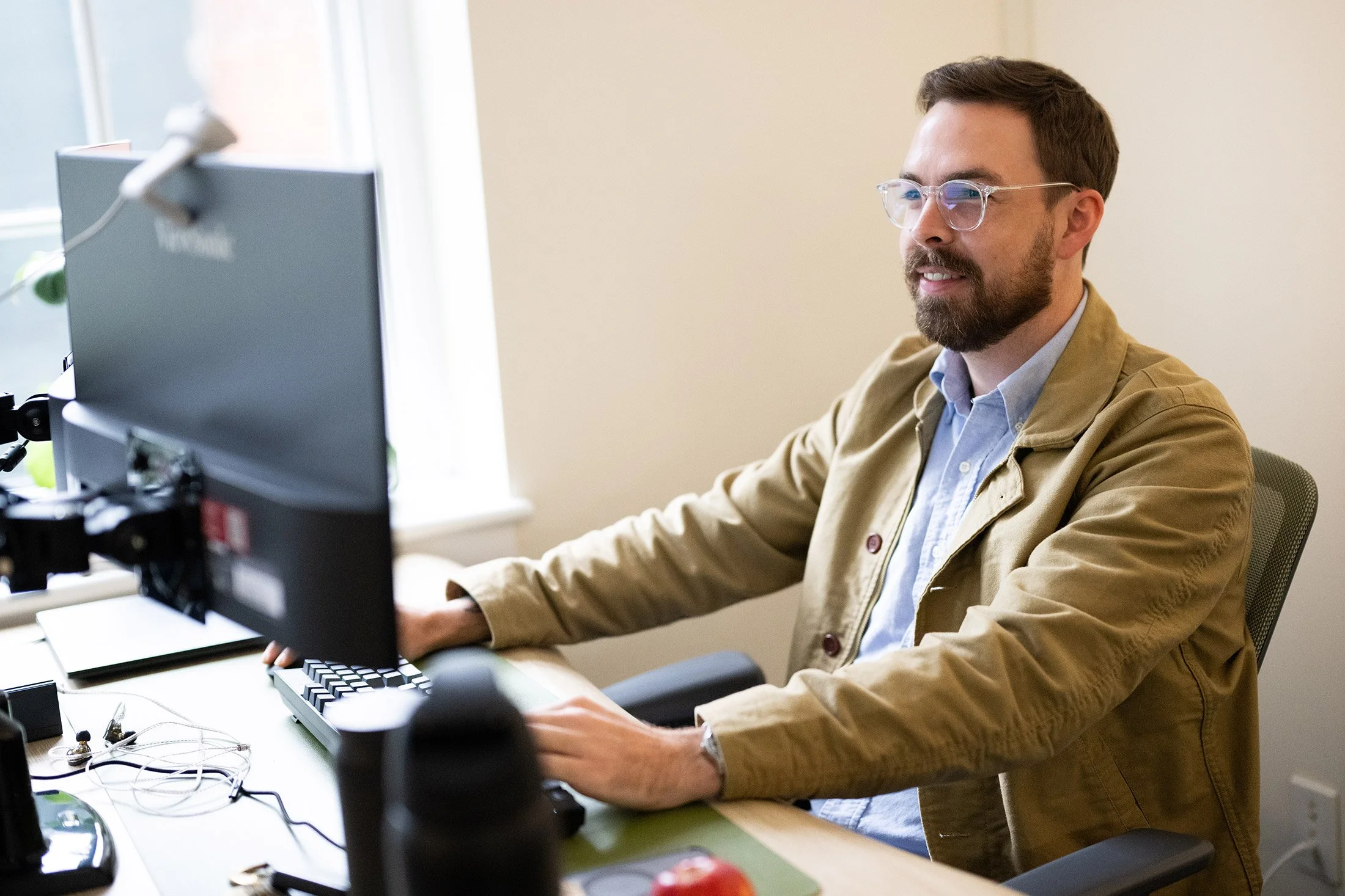 A man with glasses and a beard working at a desk with a computer monitor, keyboard, and desk accessories in a well-lit room.