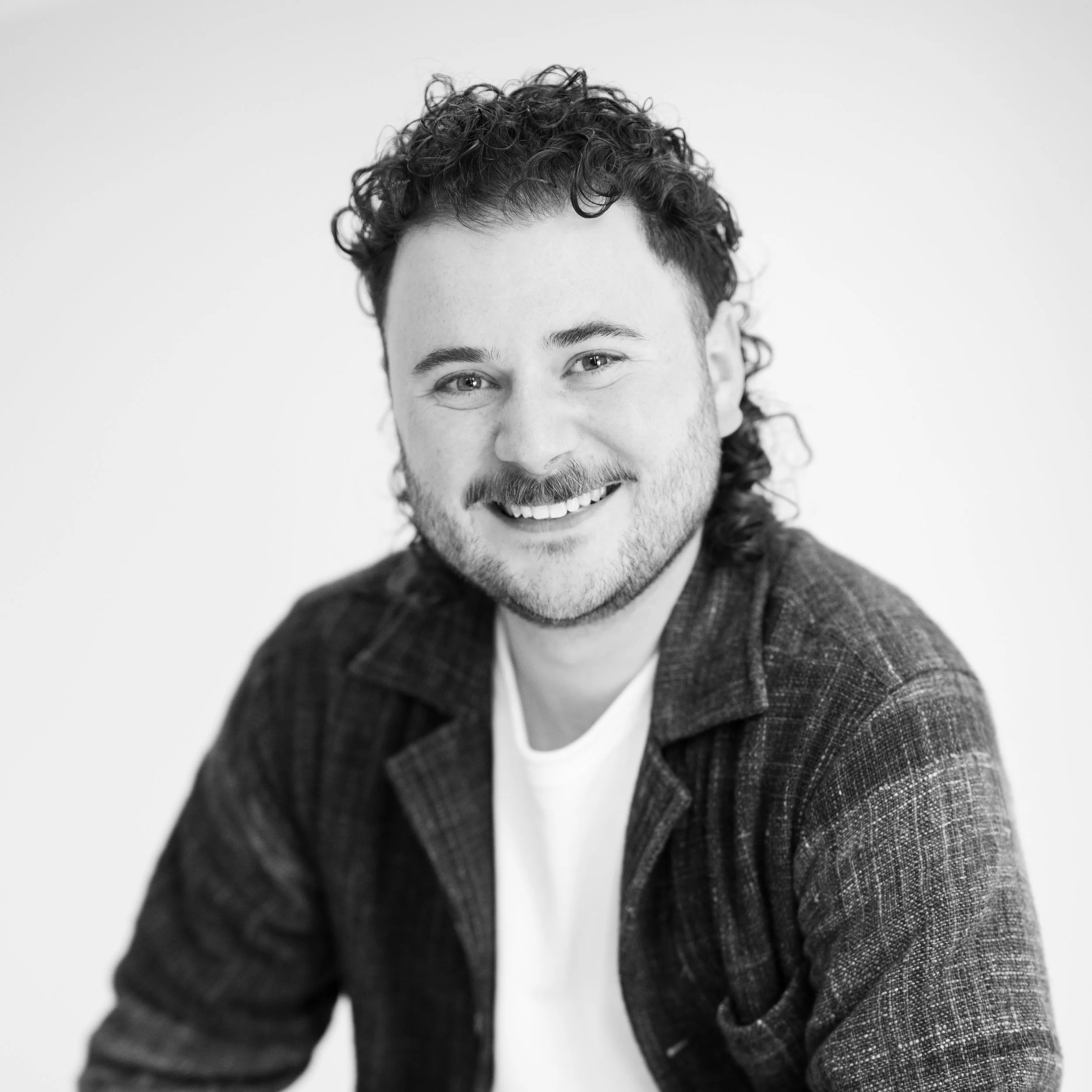 Black and white photo of a smiling man with curly hair, wearing a checked blazer and white shirt, against a plain light background.