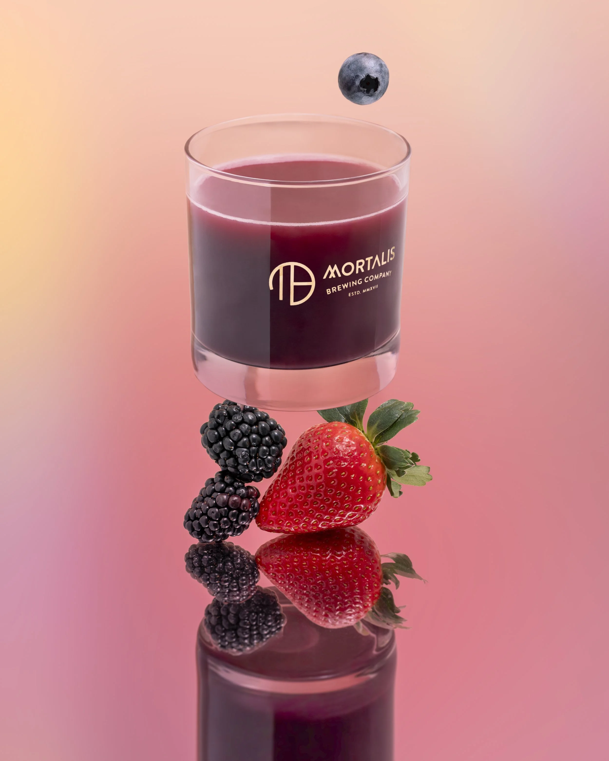 A sculptural, vertical composition featuring a strawberry and two blackberries, surreally supporting a glass of dark purple beer. Above the glass floats a single blueberry. The still life is sitting on a mirrored surface within a soft pink background