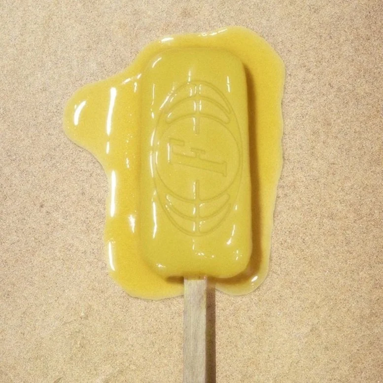 A partially melted yellow popsicle laying on the sand, viewed from above. Embossed into the popsicle is a photorealistic logo, beginning to melt.