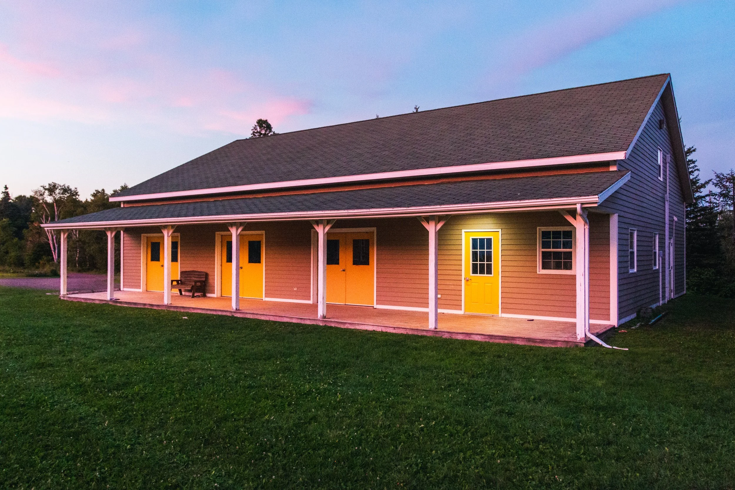 The recreation center with a sloped roof, featuring four bright yellow doors on its porch. The building is surrounded by green grass and trees, set against a pinkish-blue evening sky.