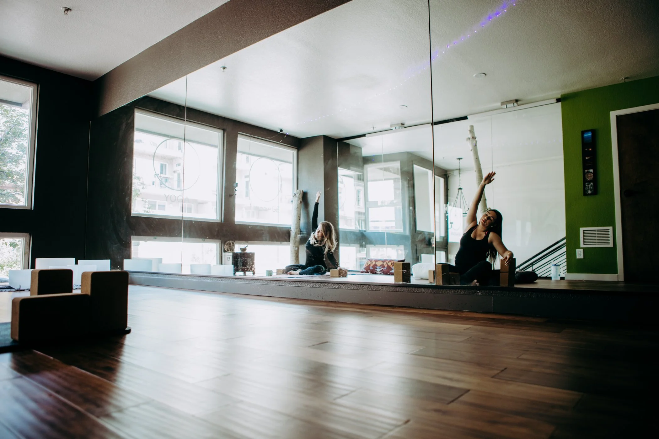Two women doing yoga stretches in a bright dance studio with large windows, mirrored wall, and wooden floors.