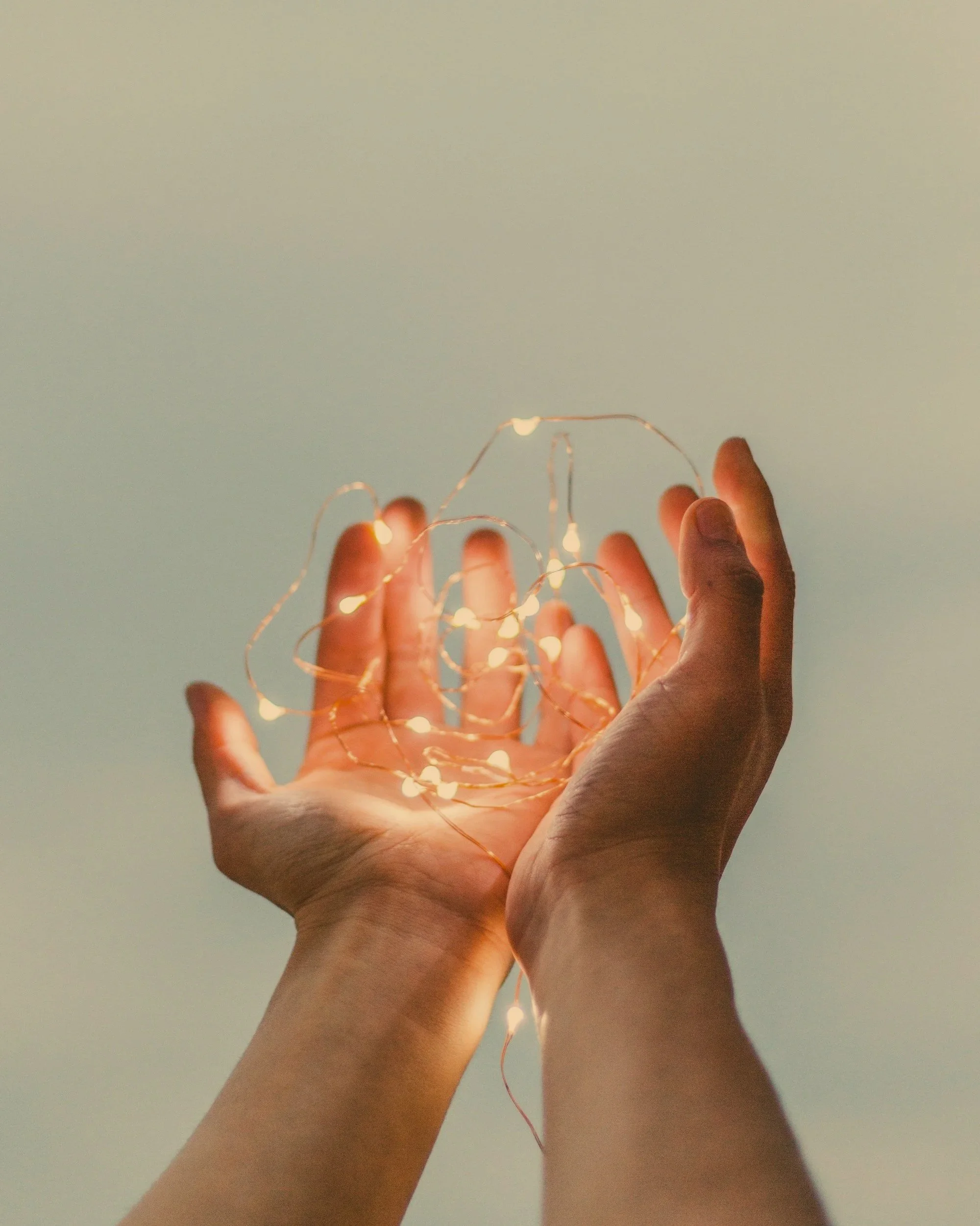 Two hands holding a string of small, glowing fairy lights against a plain wall.
