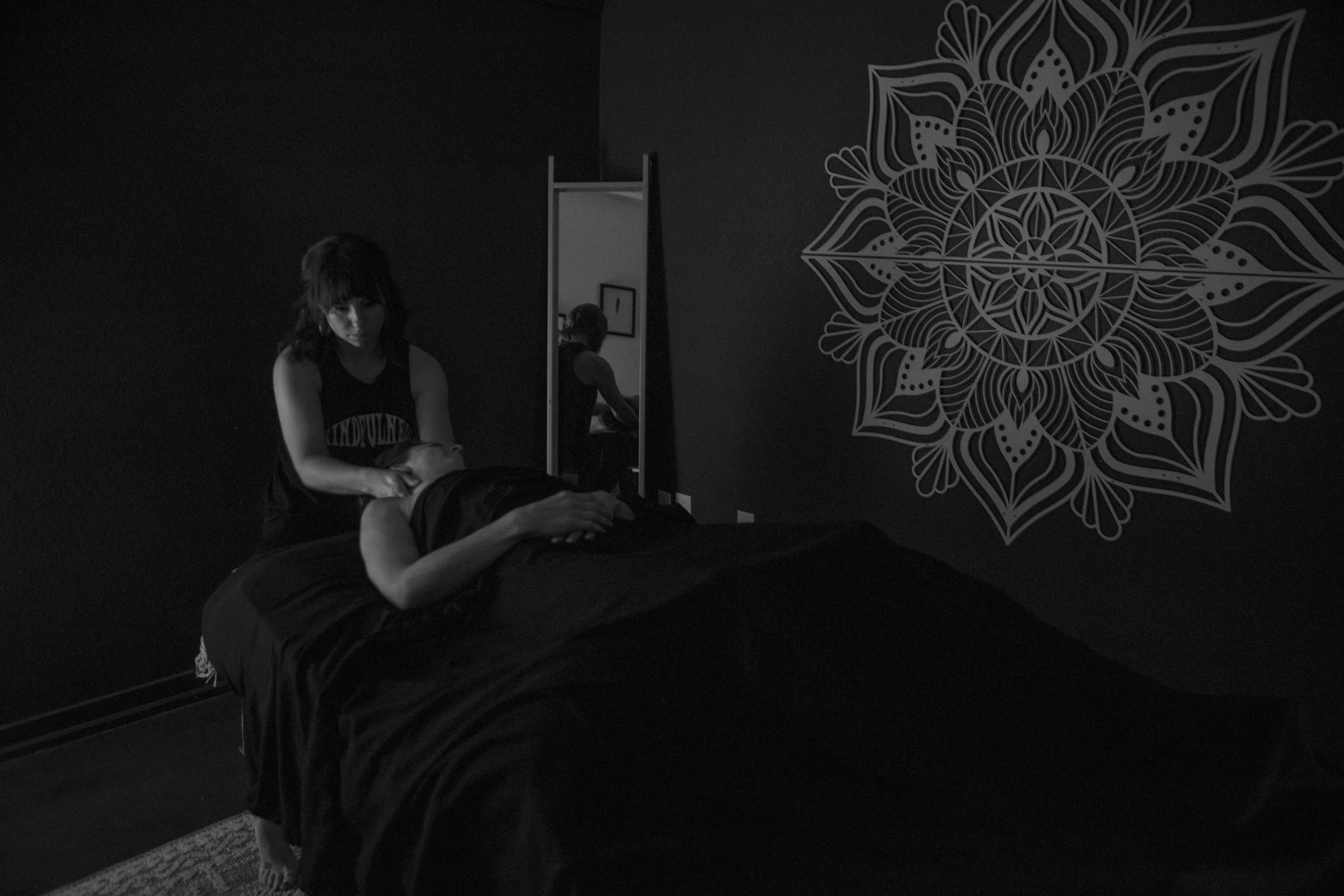 A woman is giving a massage to a person lying face up on a massage table in a dimly lit room with dark walls and a decorative mandala artwork on the wall.