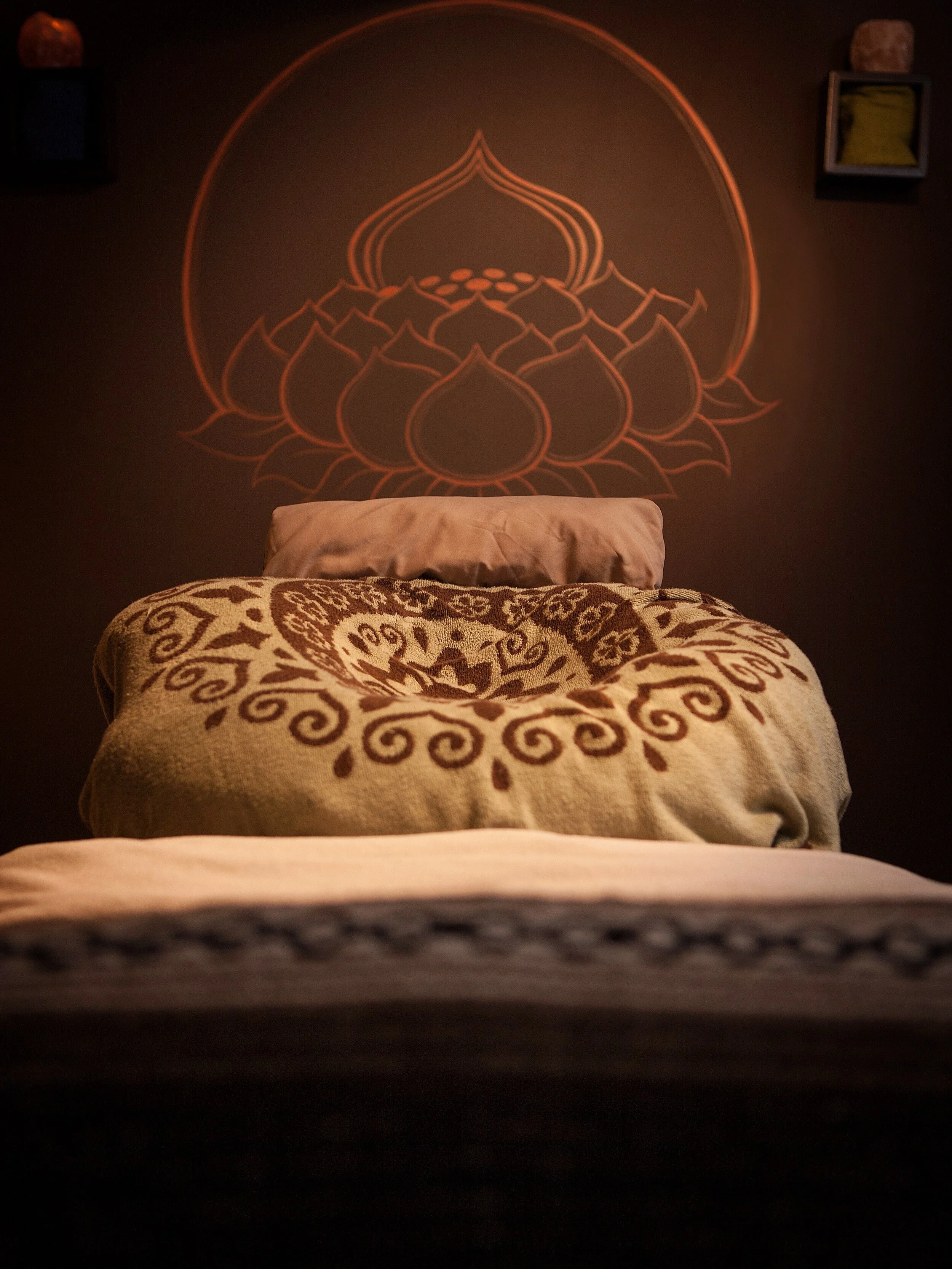 A neatly made bed with patterned blankets and a pillow, with a glowing orange lotus and decorative design projected on the dark wall above the headboard.