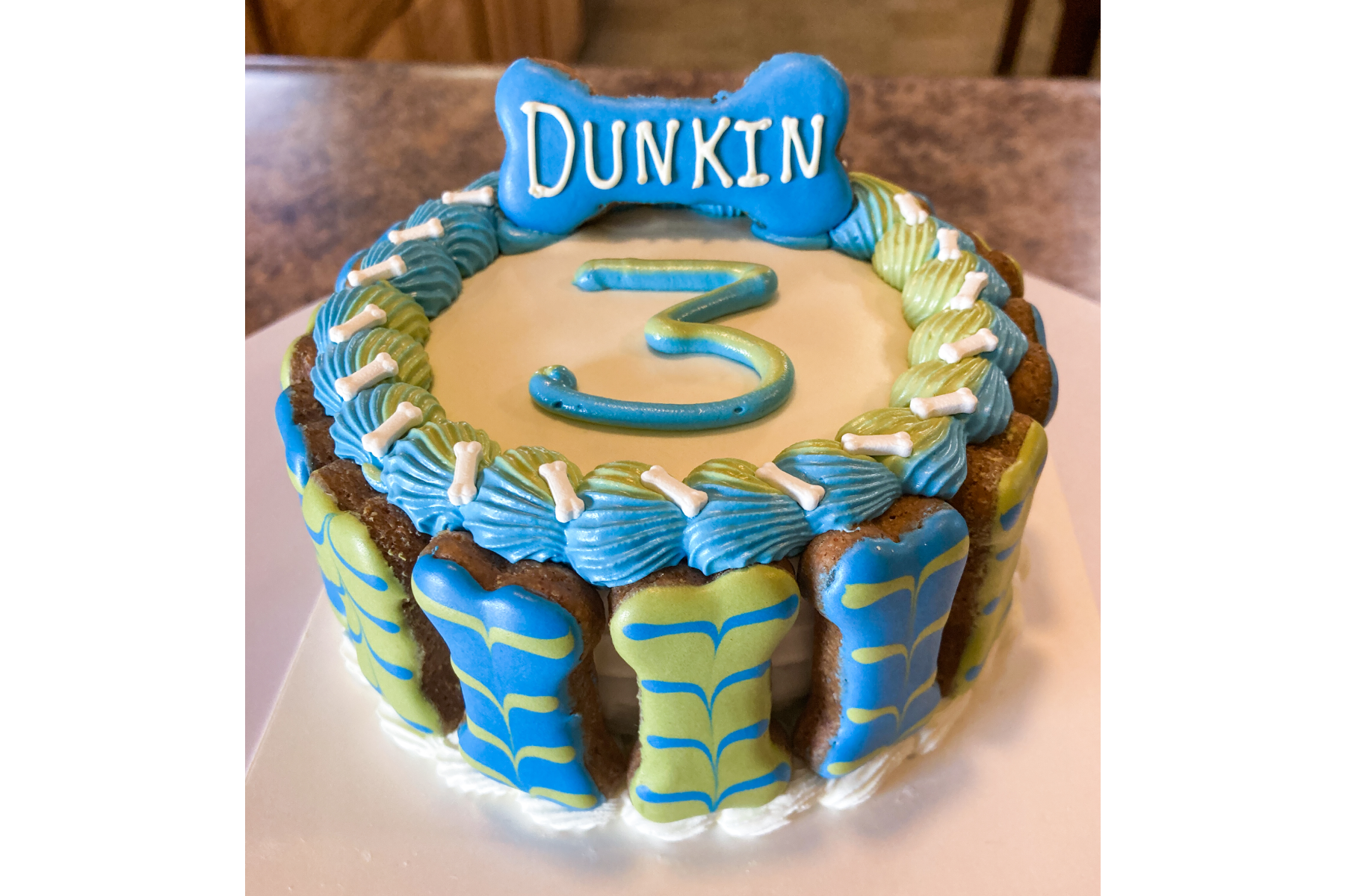Image of Birthday / Gotcha Day Cake. 2-layer cake, bones around the sides, age and bone with pup's name on top. Iced in white and decorated in blue and green.