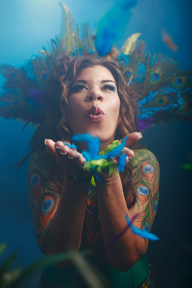 edmonton fantasy boudoir studio portrait woman with peacock bodypaint blows colorful feathers playfully smiling