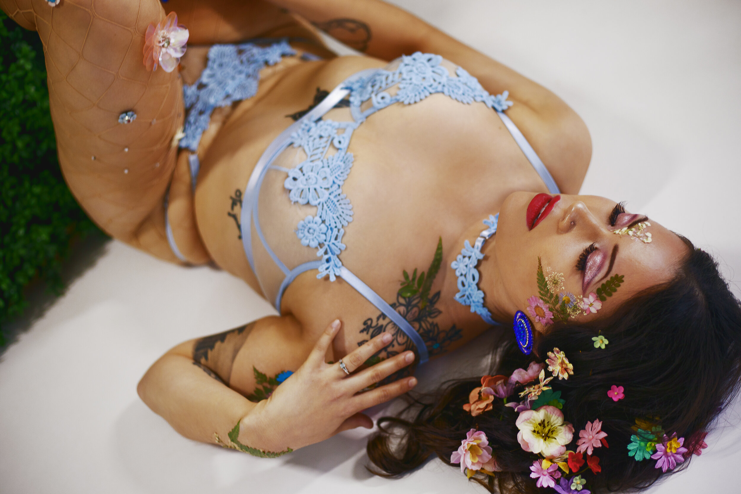 edmonton bipoc boutique boudoir portrait woman laying against plant wall adorned with flowers in hair and blue lace lingerie