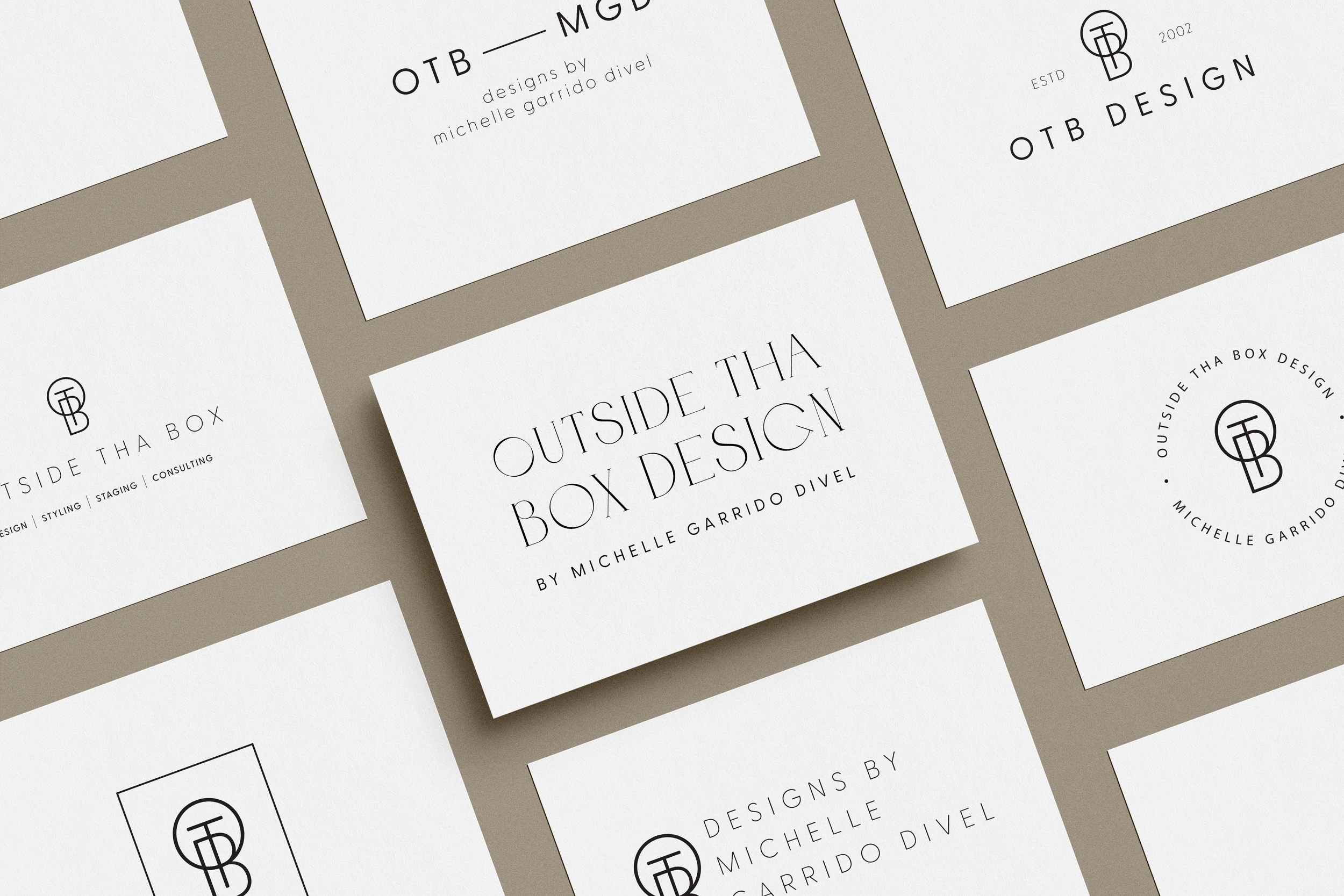 Multiple white business cards featuring calligraphy and line art logos for "Outside Tha Box Design" and "OTB Design" by Michelle Garrido Divel, arranged on a beige surface.