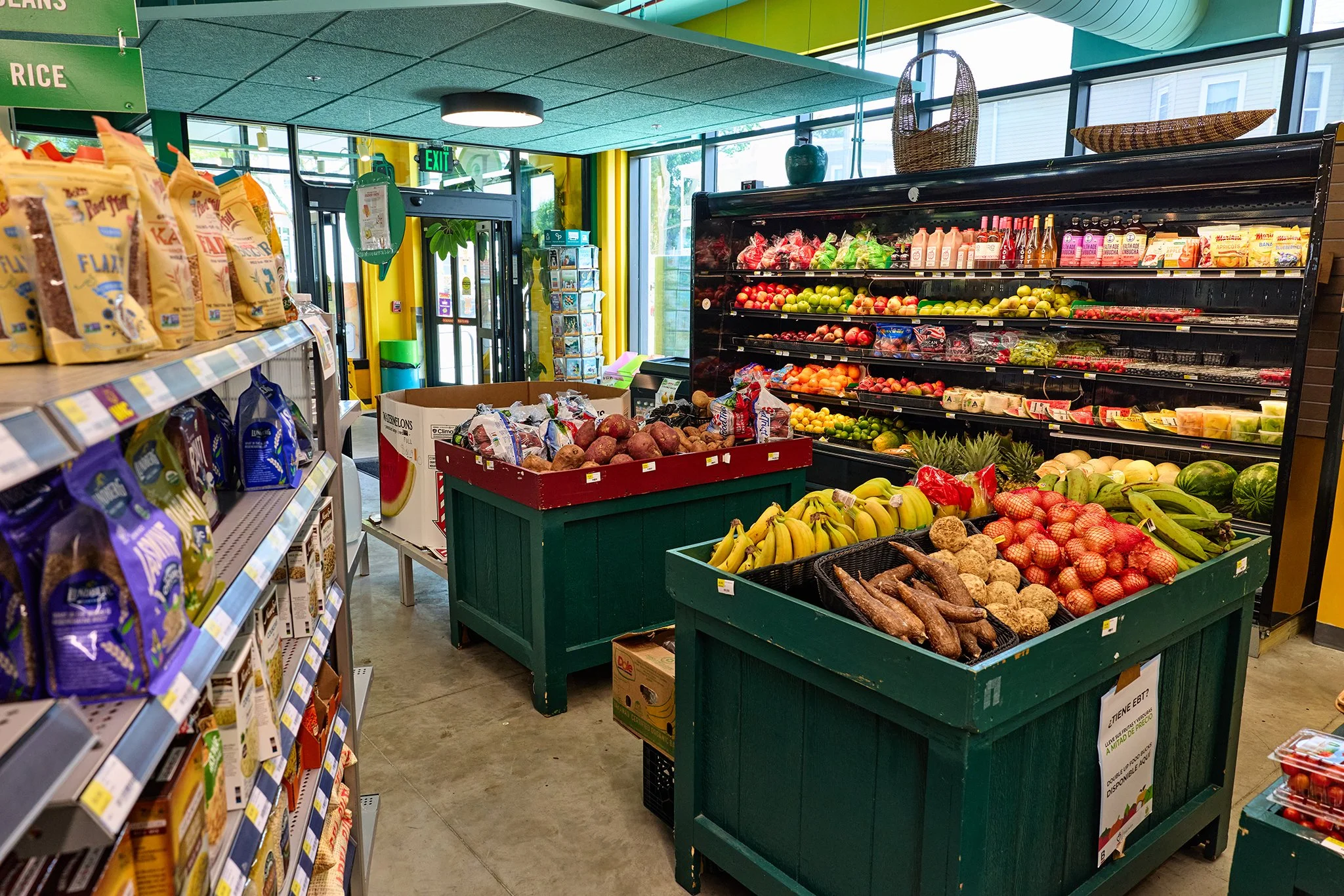 Inside a grocery store produce section with green crates filled with bananas, sweet potatoes, onions, and other fruits and vegetables. To the left is a shelf with snack packets, and in the background, there are glass doors, a yellow wall, and large windows letting in natural light.