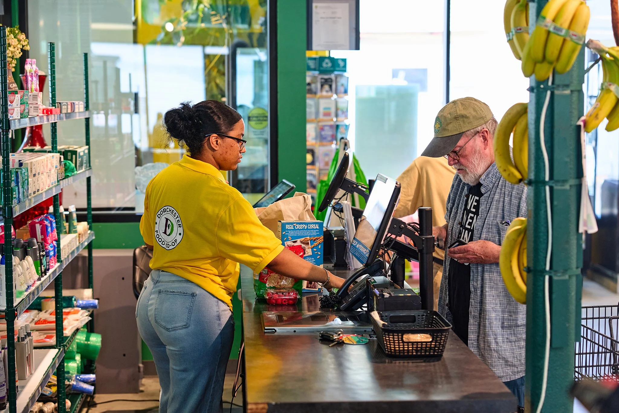 A woman wearing a yellow shirt and glasses is working at a checkout counter with a customer in a grocery store. The customer, an older man with glasses, a beard, and a cap, is handing over payment. Items like cereal and bananas are on the counter, with shelves of products in the background.