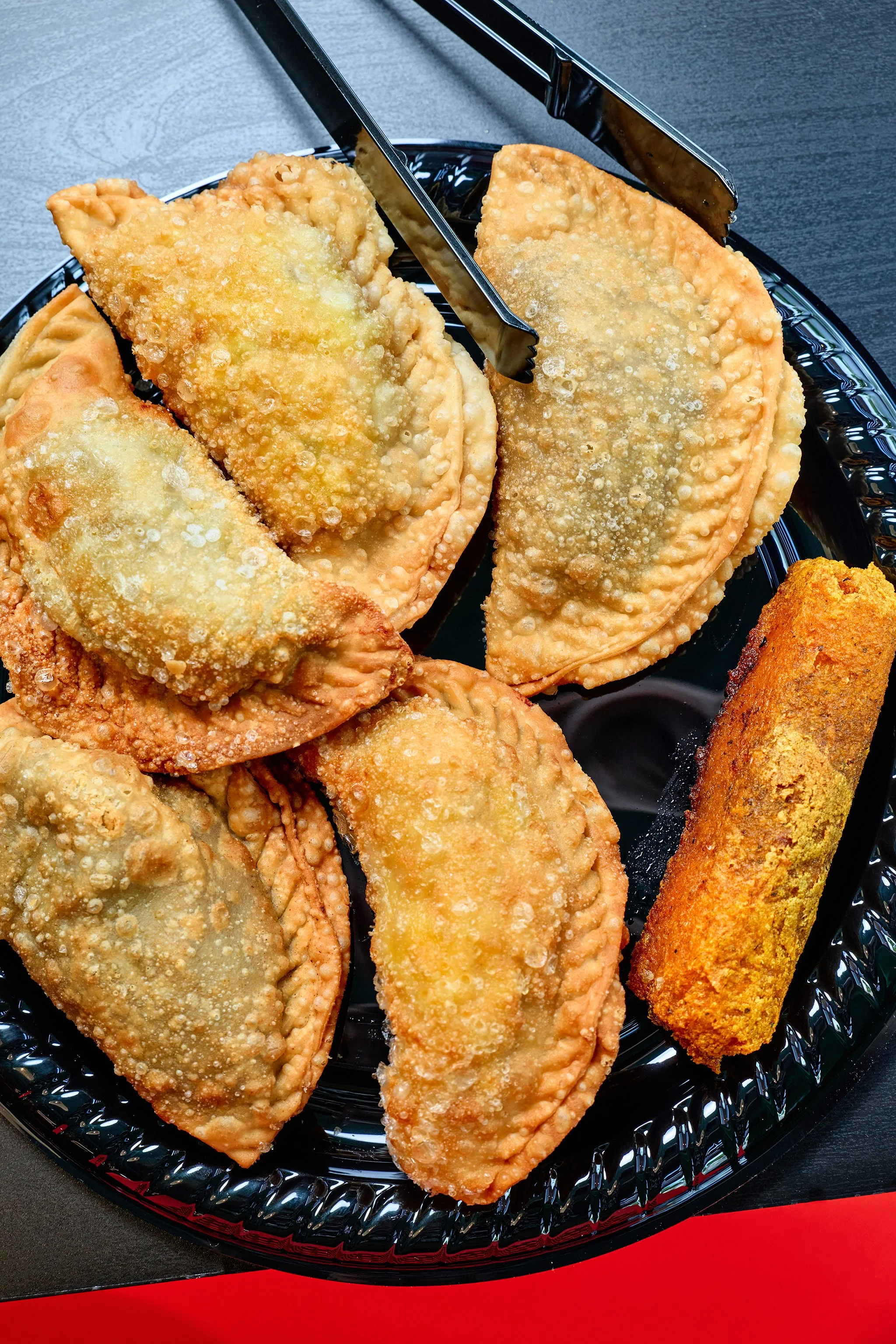 Close-up of fried food items on a black plastic tray, including fried fish fillets and a fried roll, on a red and black surface.